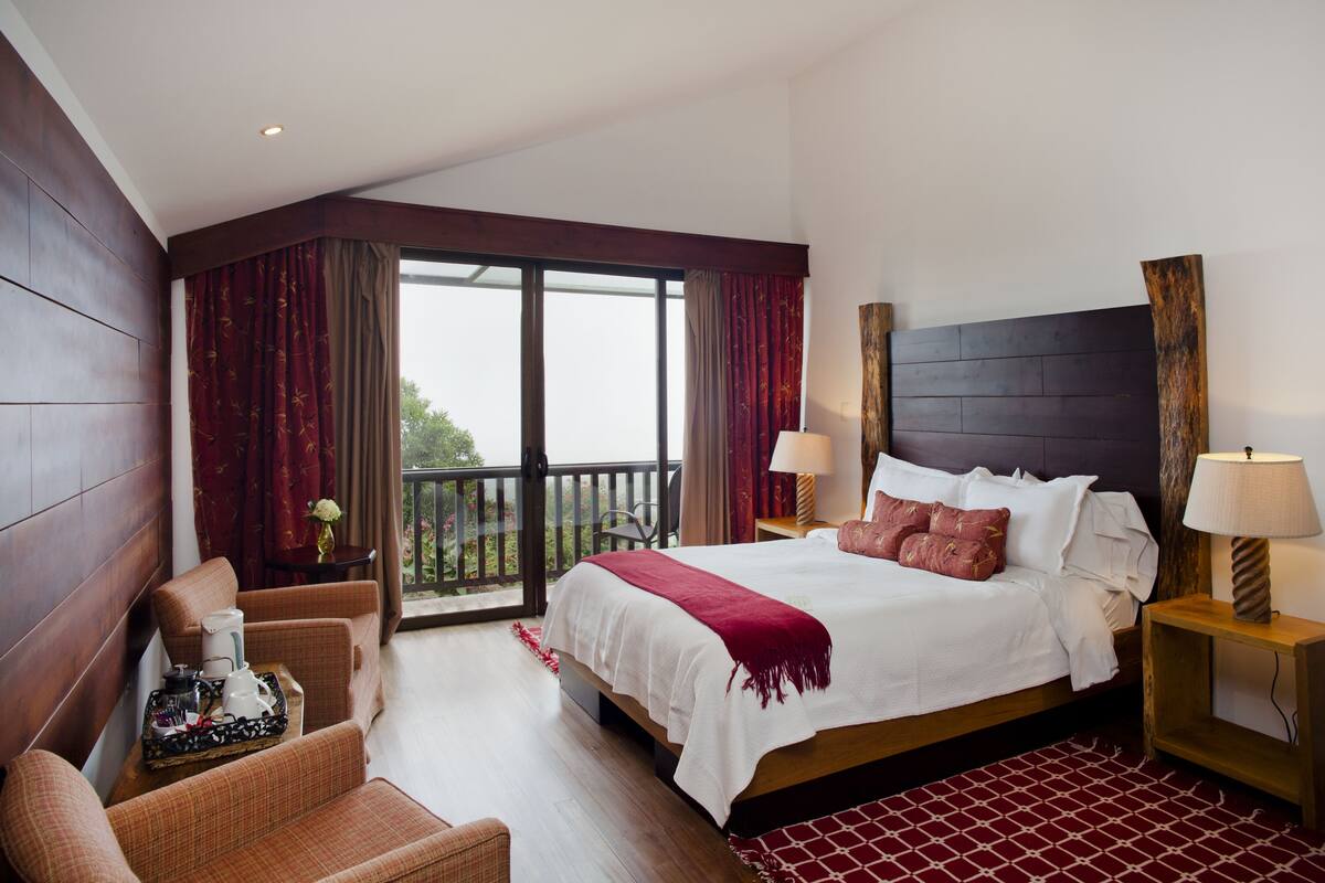 Habitación de hotel con una cama grande, dos mesitas de noche con lámparas, un cabecero de madera, un balcón con cortinas y una zona de estar con una mesa y sillas.