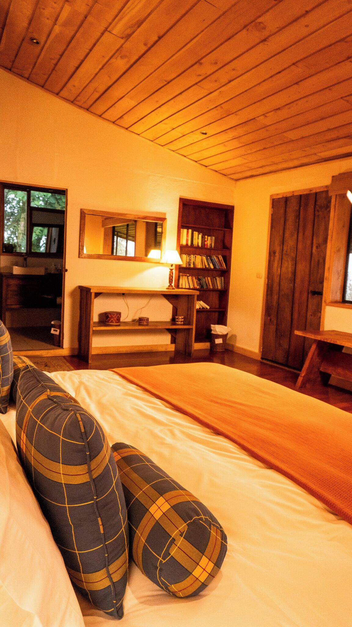 A cozy bedroom with a wooden ceiling, a bed with plaid pillows, a bedside table with a lamp, a bookshelf, and a mirror.