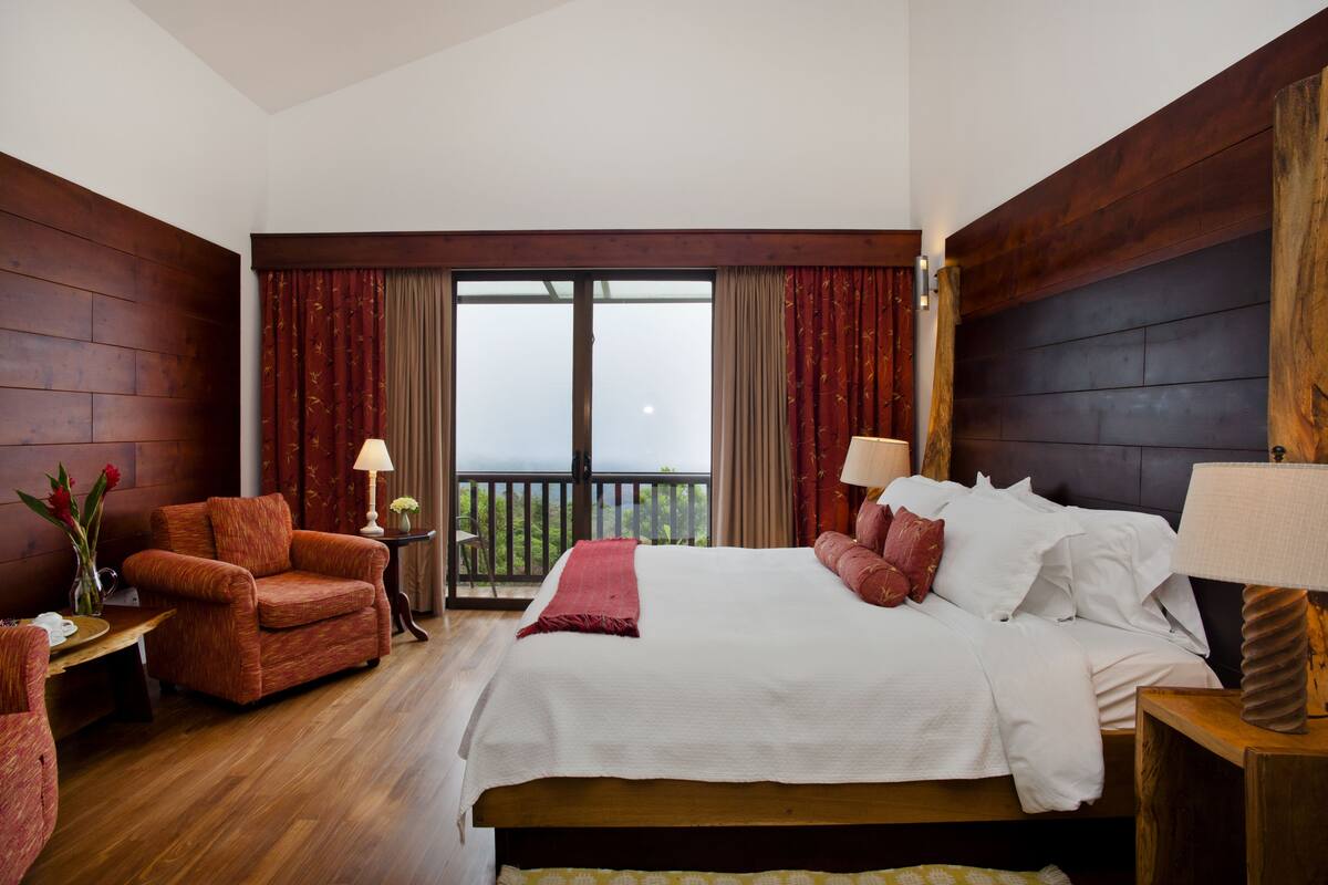 Una habitación de hotel con una cama grande, una zona de estar con una silla, una mesita y un balcón con cortinas.