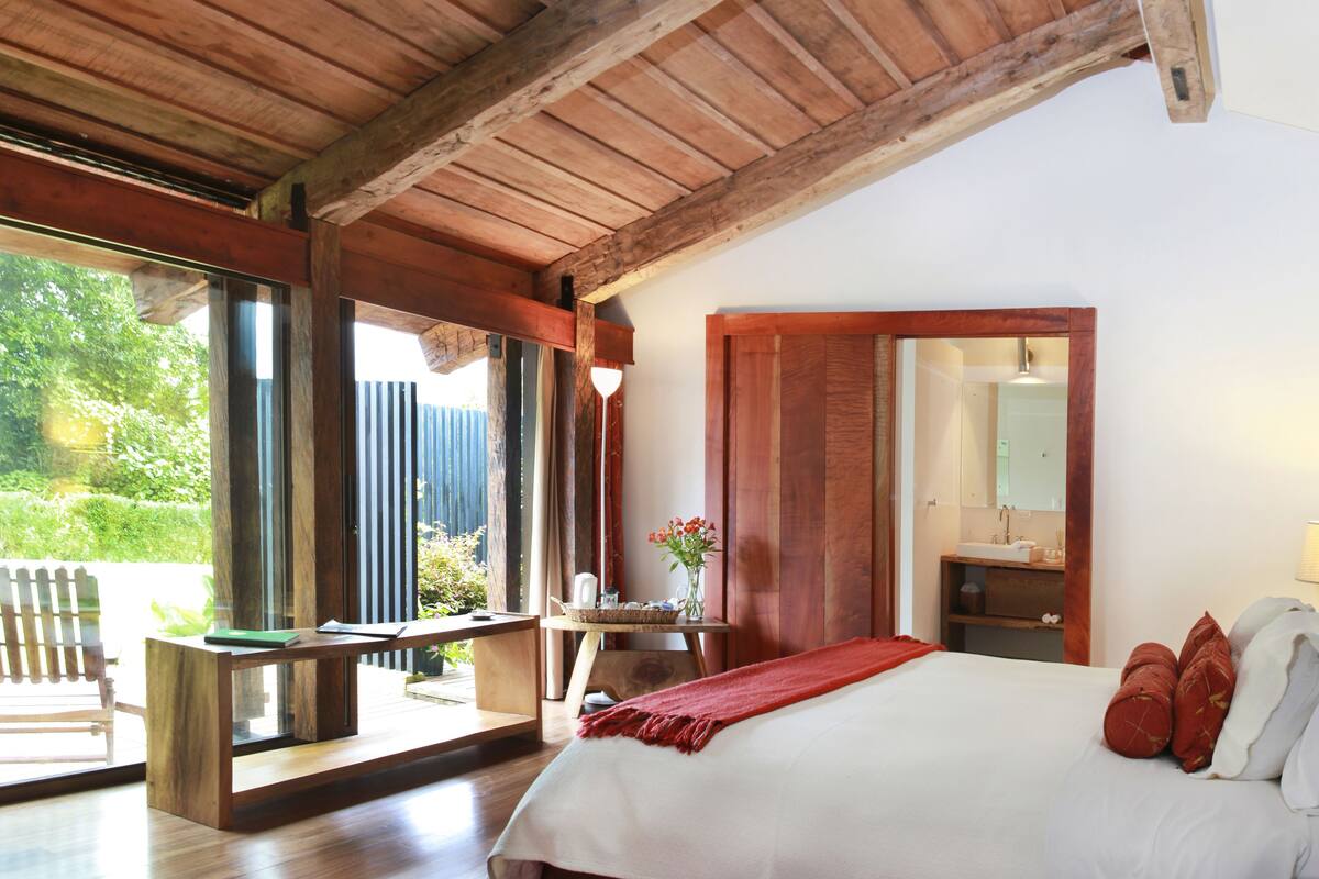 Un dormitorio con una cama grande, vigas de madera en el techo, un ventanal con vistas al verdor y una pequeña mesa con un florero.