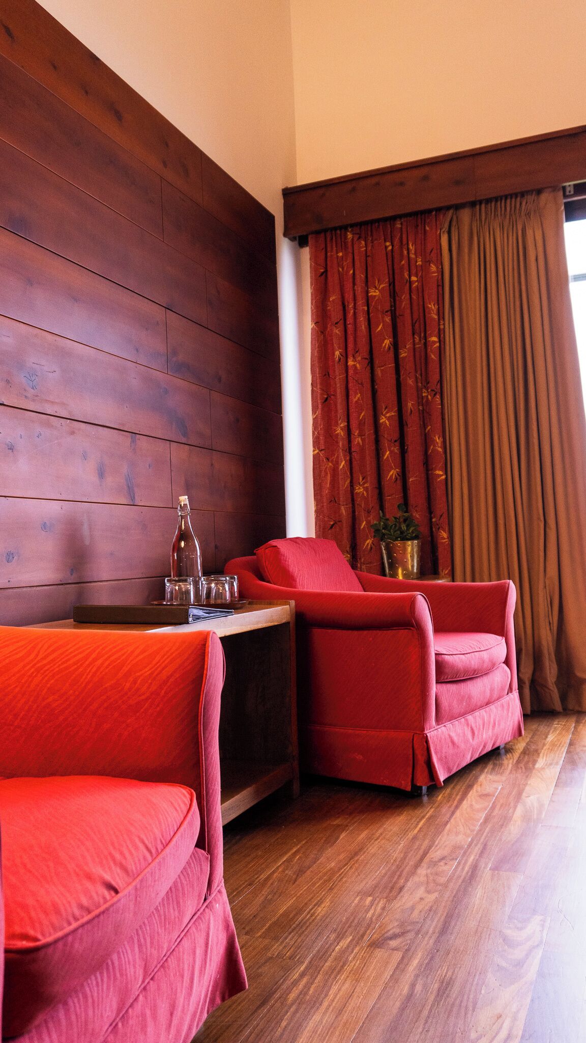 A room with wooden panel walls, a red sofa, a red armchair, a small table with a bottle, glasses, and a plant.