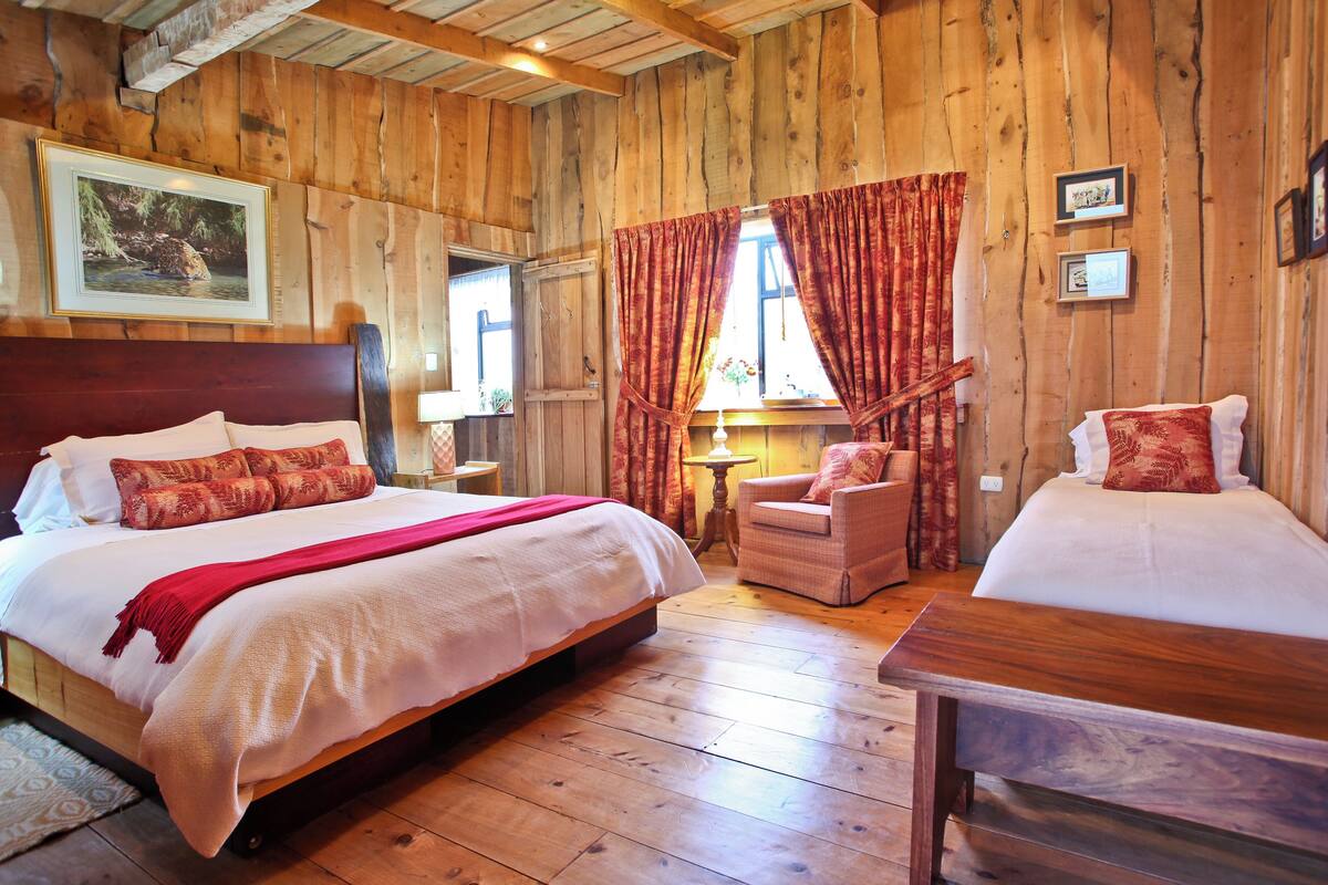 Un dormitorio estilo cabaña de madera con dos camas, cabecero de madera, mesita de noche, una silla y una ventana con cortinas.