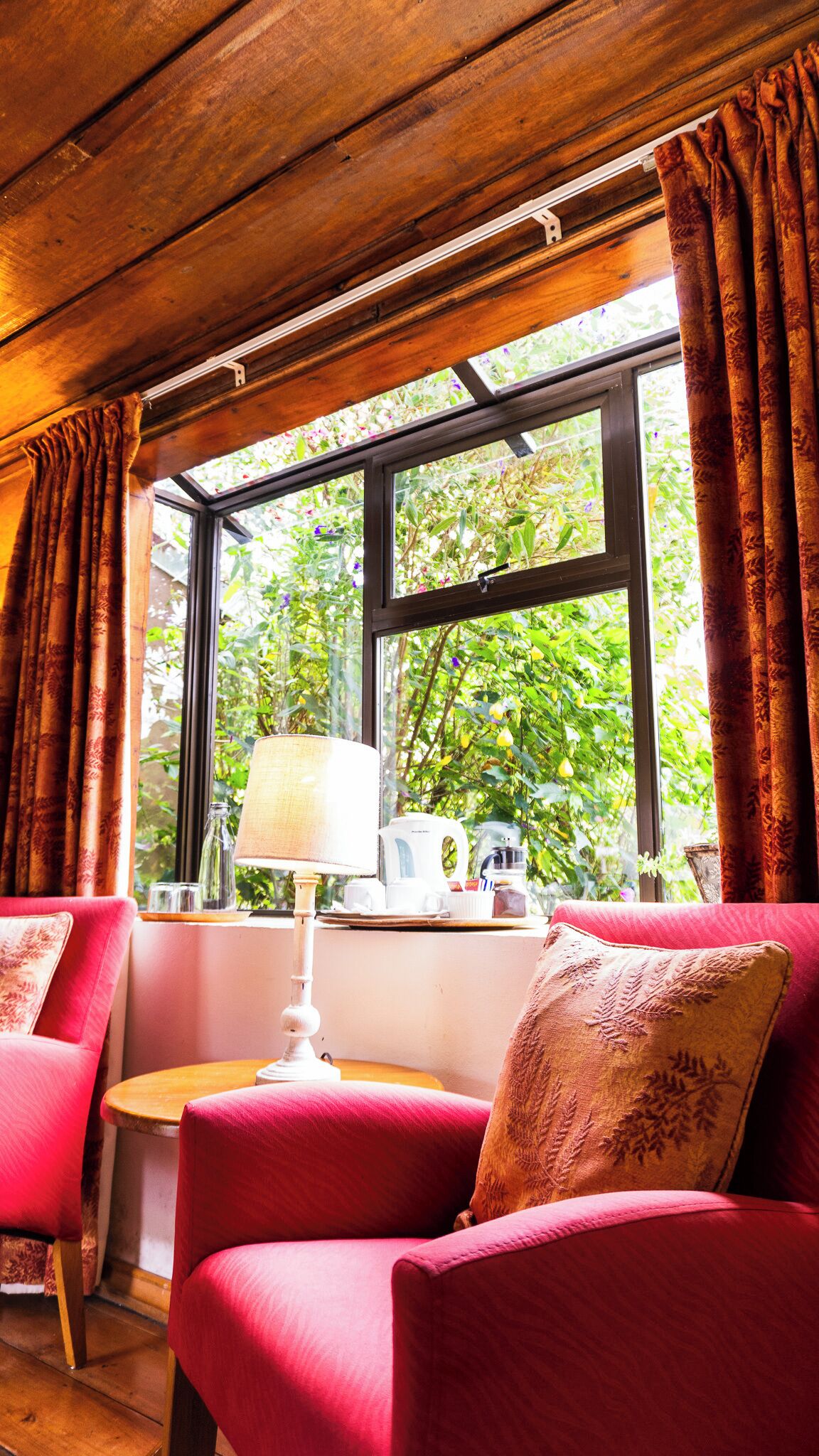 A cozy room with a wooden ceiling, a window with curtains, a table with a lamp, and two red sofas with patterned cushions.