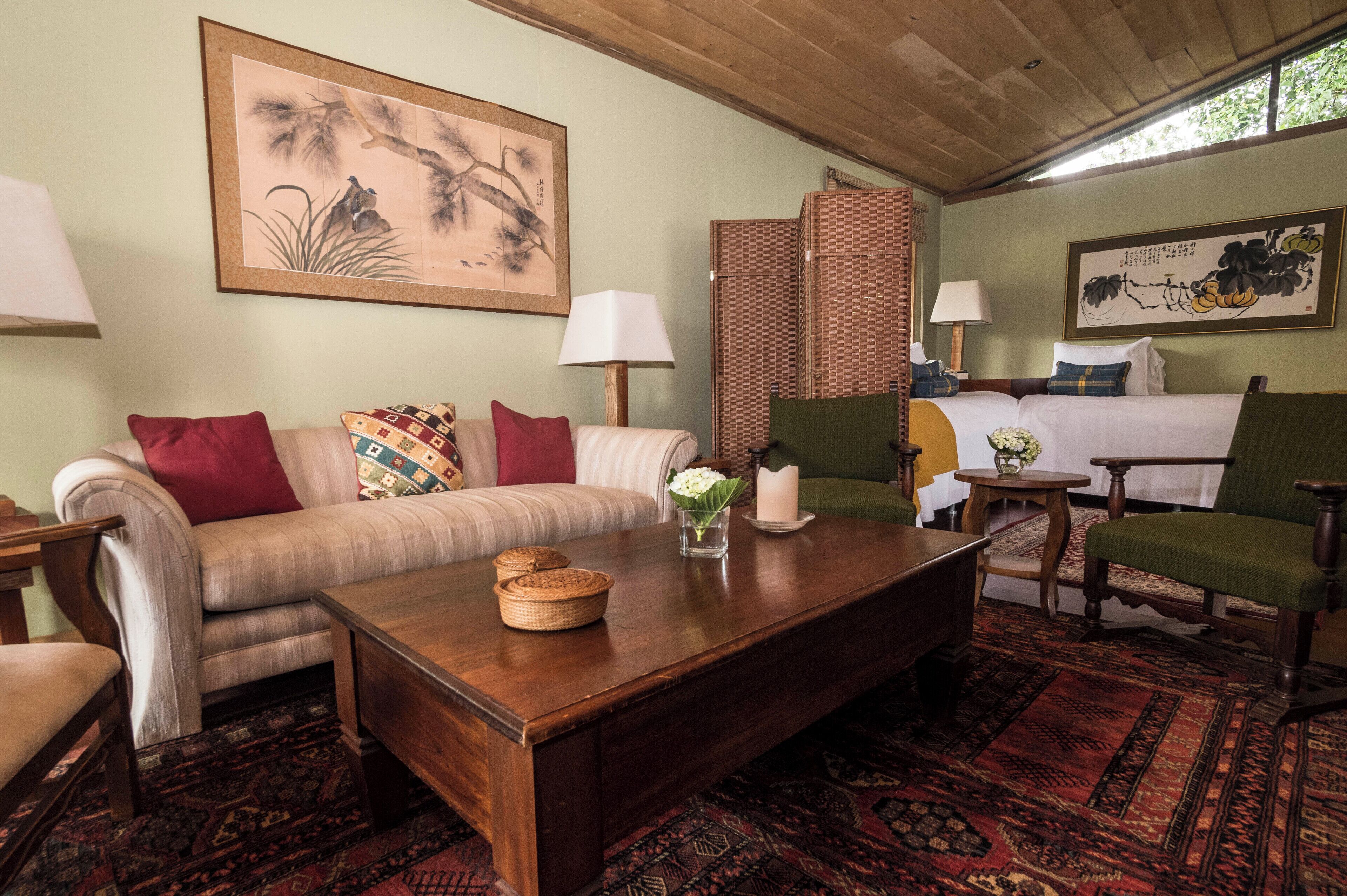 A room with a sofa, a wooden coffee table, a green armchair, a bed with a green chair, and a large painting on the wall.