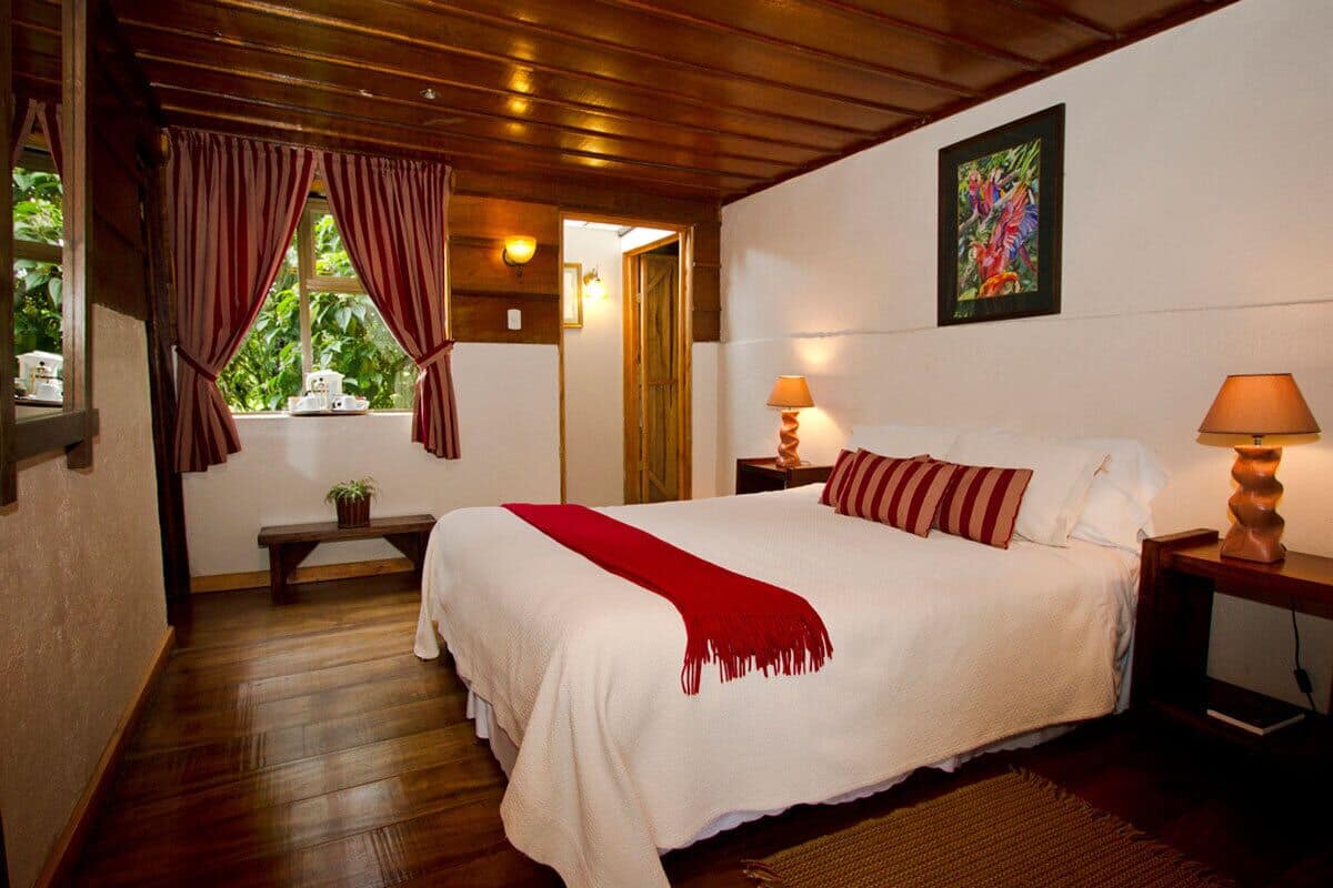 Un dormitorio con una cama grande, suelo de madera, un banco, una ventana con cortinas y un cuadro en la pared.