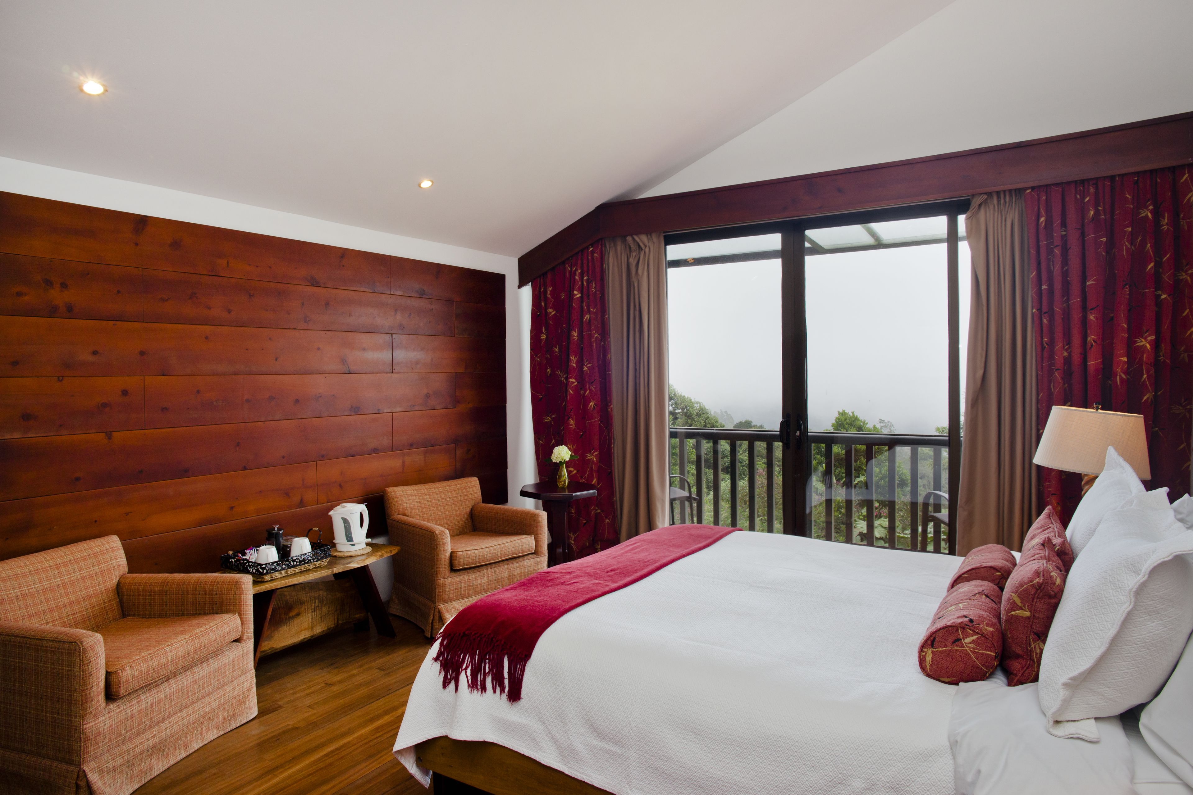 Habitación de hotel con pared de madera, una cama grande con almohadas rojas, una mesa de madera con un hervidor, una silla y vistas al exterior a través de un ventanal.