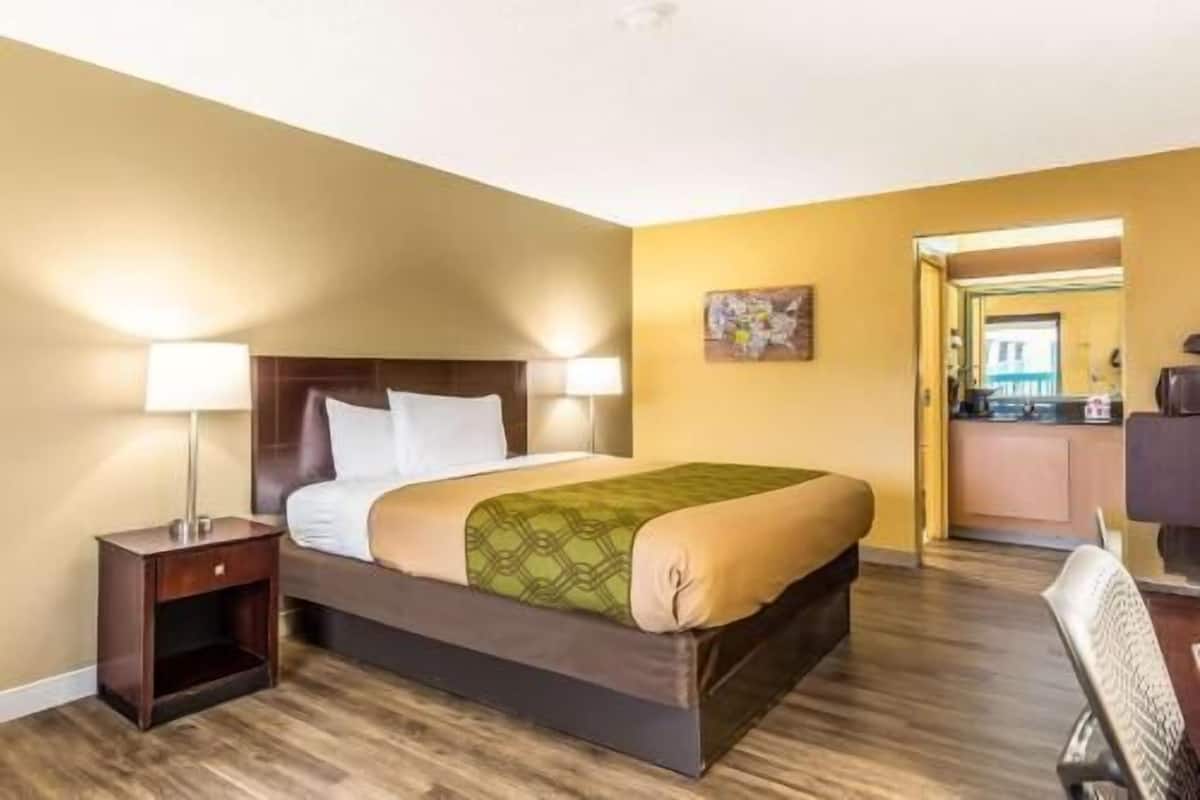 A hotel room with a bed, bedside lamps, a small wooden nightstand, a green patterned bedspread, a painting on the wall, and a view of the kitchen through an open door.