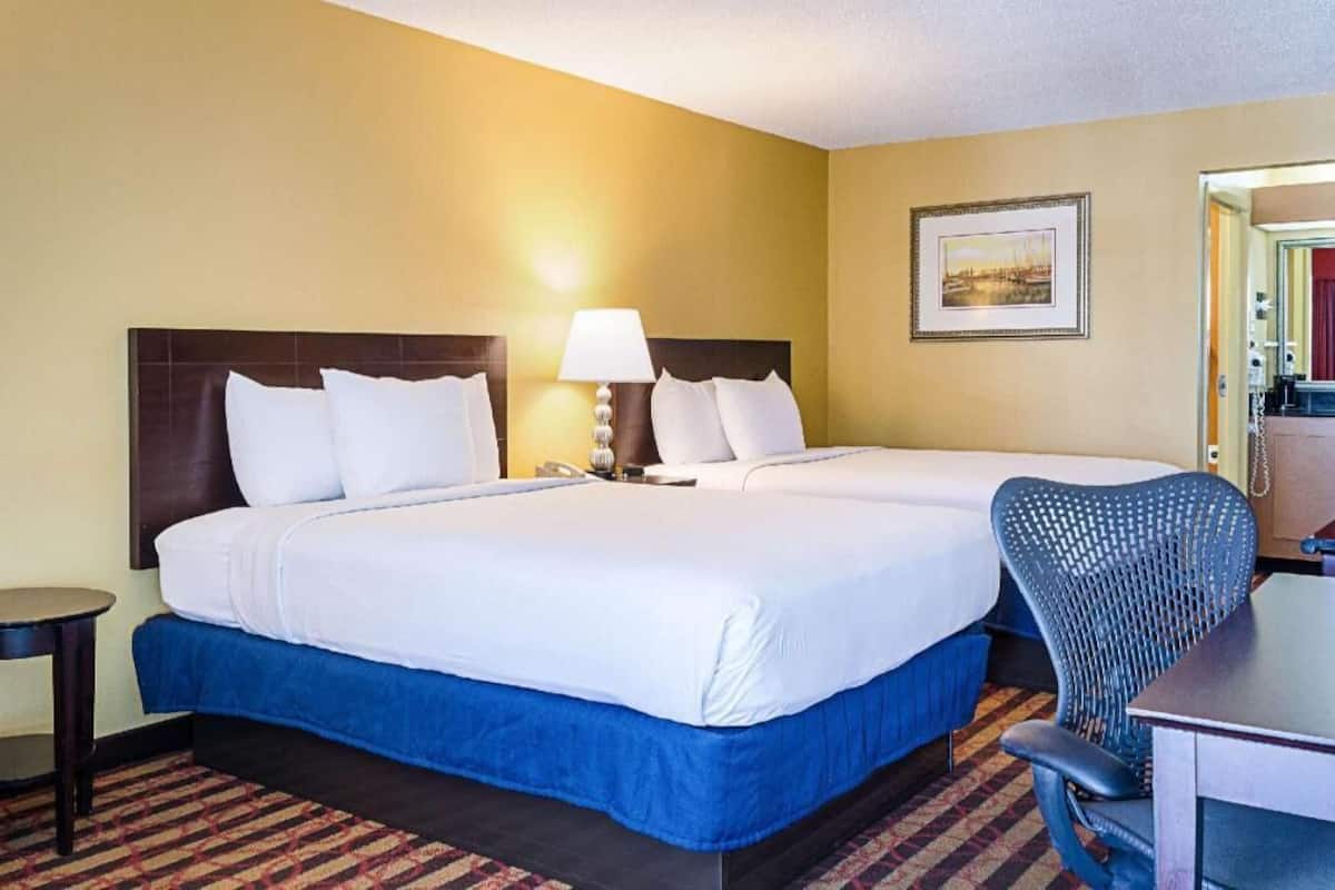 A hotel room with two beds, a desk, a chair, a lamp, and a framed picture on the wall.