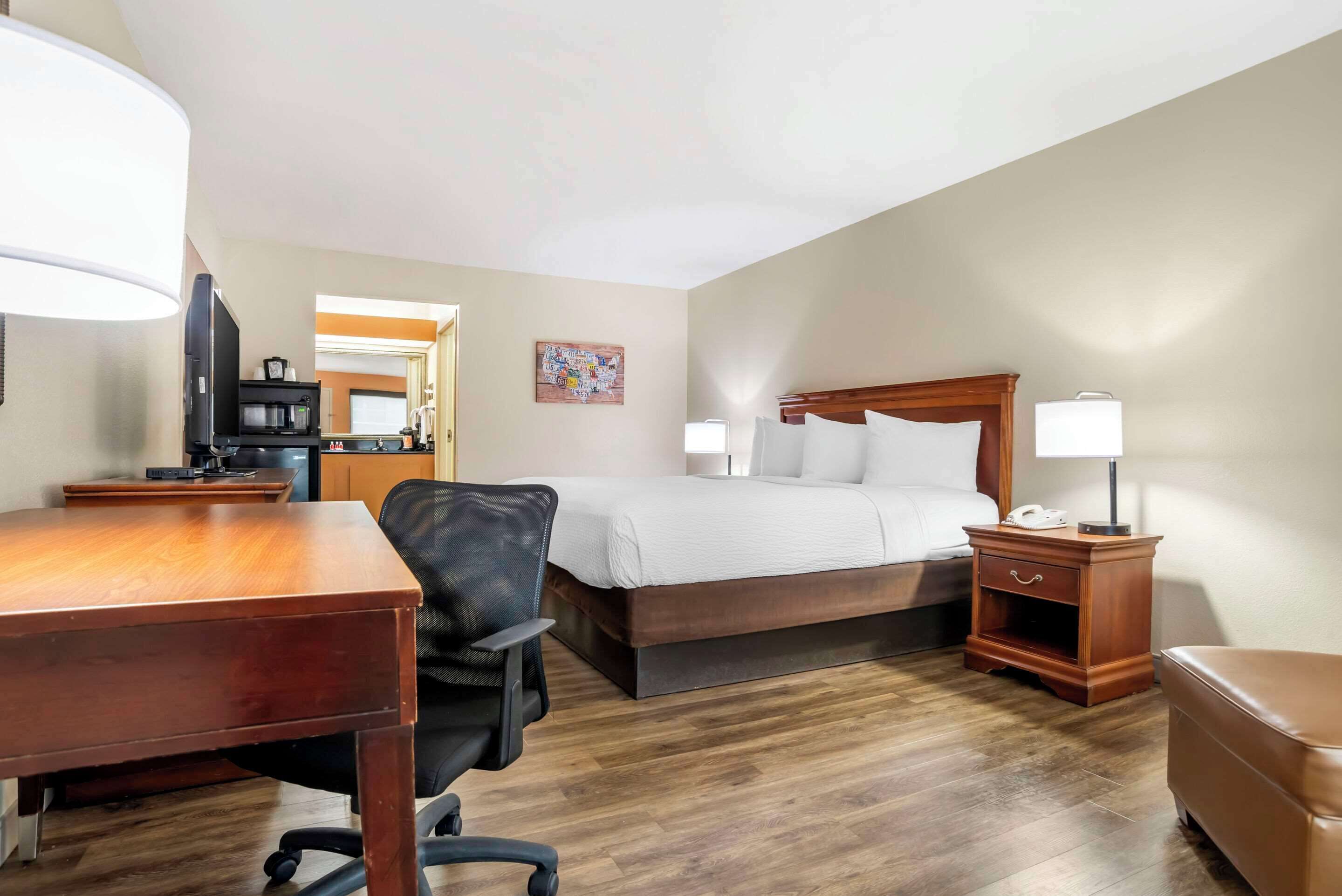 A hotel room with a wooden desk, a black office chair, a bed with white bedding, a nightstand with a lamp, and a wooden floor.