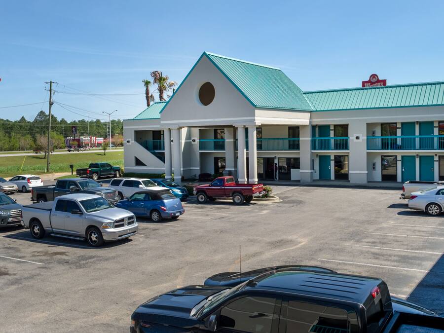 A hotel with a green roof and white exterior, parking lot with various vehicles, and a flagpole with an American flag.