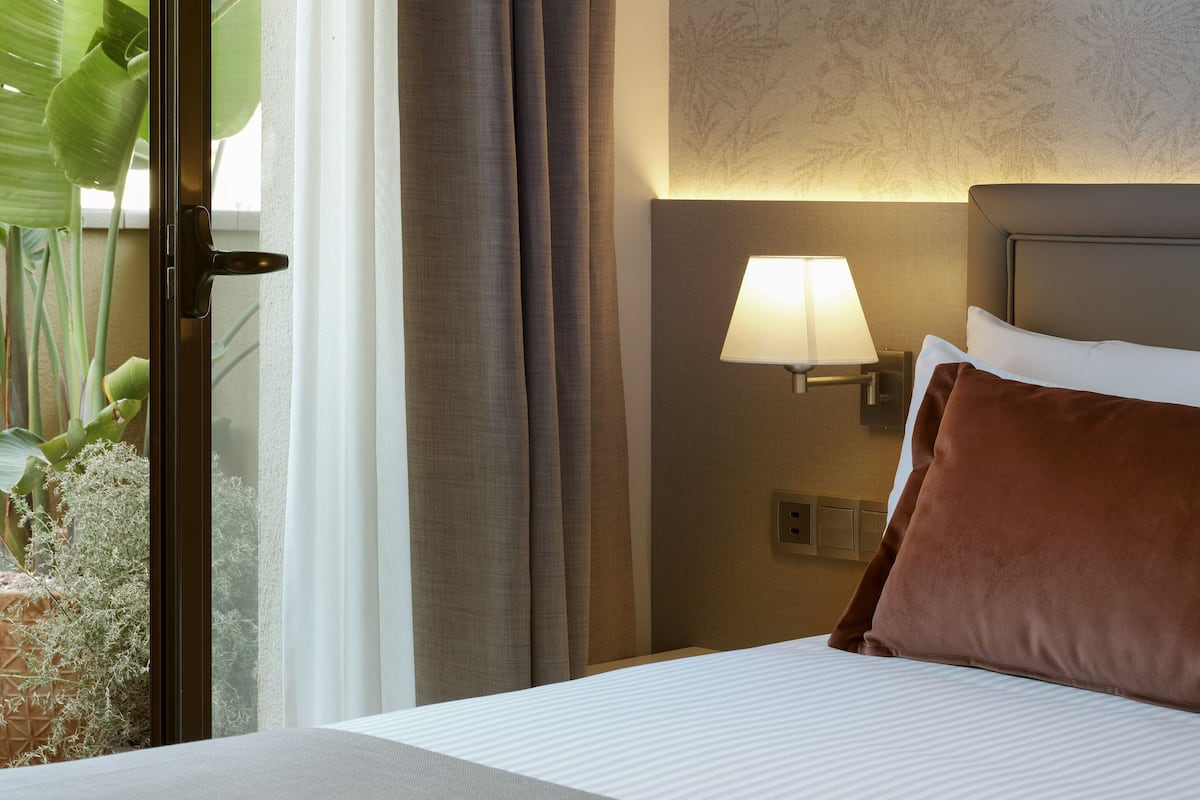 Una habitación de hotel con cama, una mesita de noche con lámpara, una ventana con cortinas y una planta afuera.