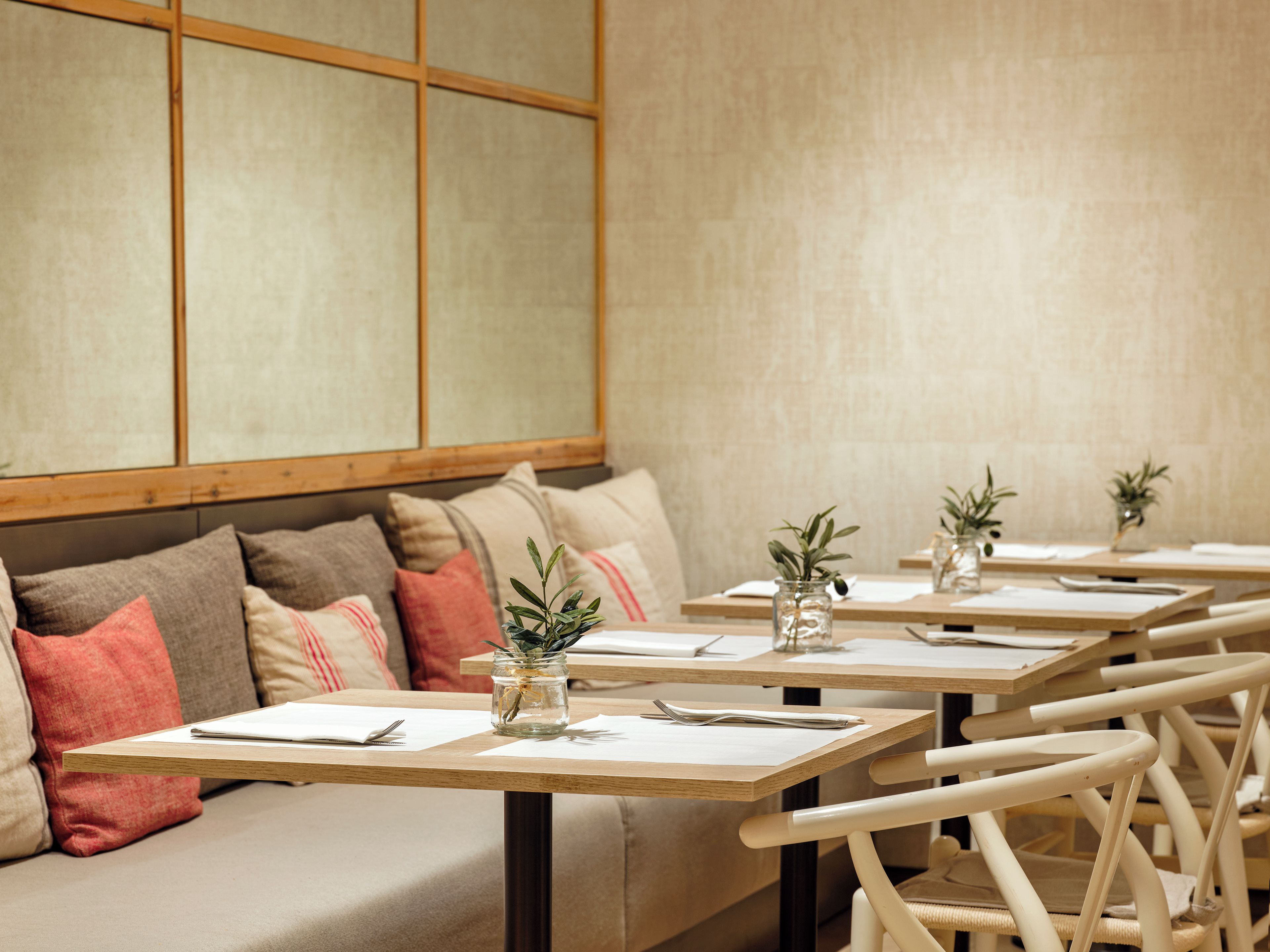 Et restaurantinteriør med flere borde, alle dækket med en bordskåner, gaffel og kniv. Der står plantekasser på bordene, og i baggrunden ses en sofa med hynder.