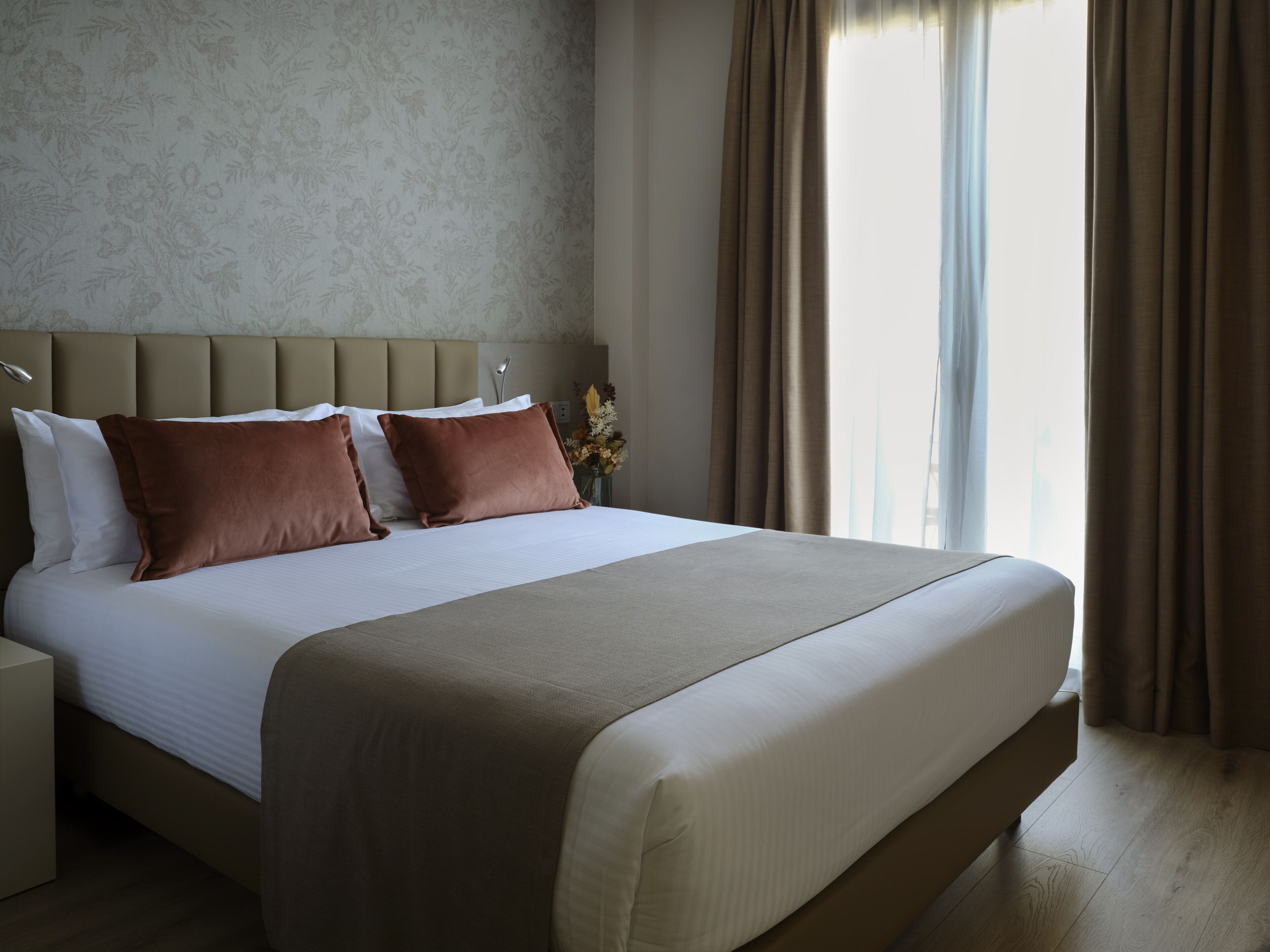 Un letto rifatto con coperte ordinate, una testata, un comodino e un vaso con fiori.