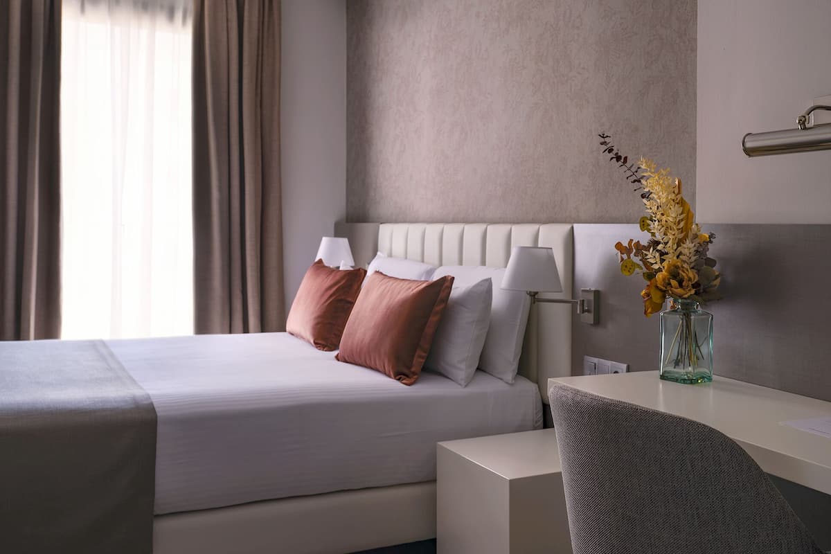 Una habitación de hotel moderna con una cama bien hecha, una mesita de noche con lámpara y un jarrón con flores.
