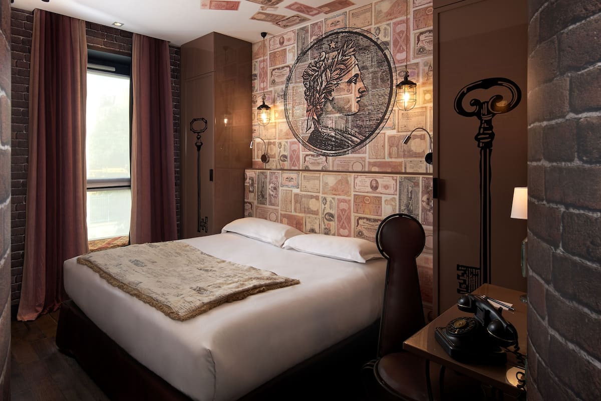 Quarto de hotel com parede de tijolos, uma cama grande com edredom branco, mesa de cabeceira de madeira e luminária fixada na parede.