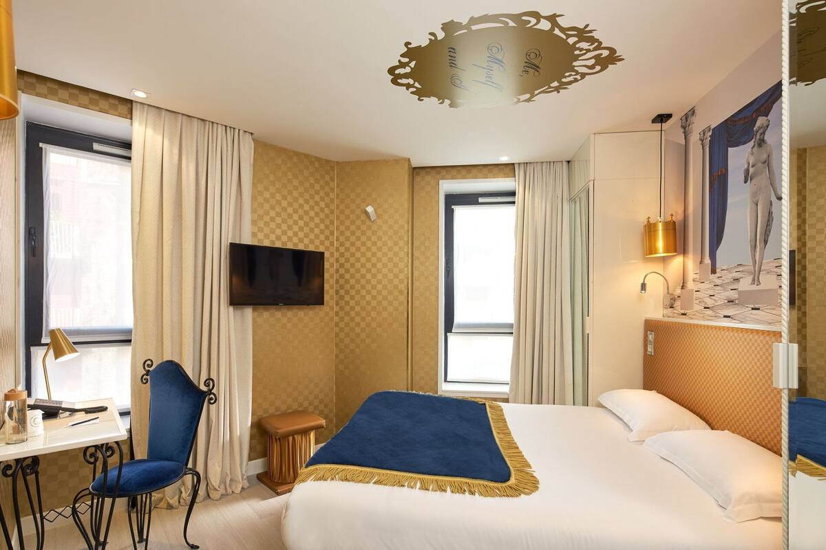 Quarto de hotel com cama, escrivaninha com cadeira, televisão e janela com cortinas.