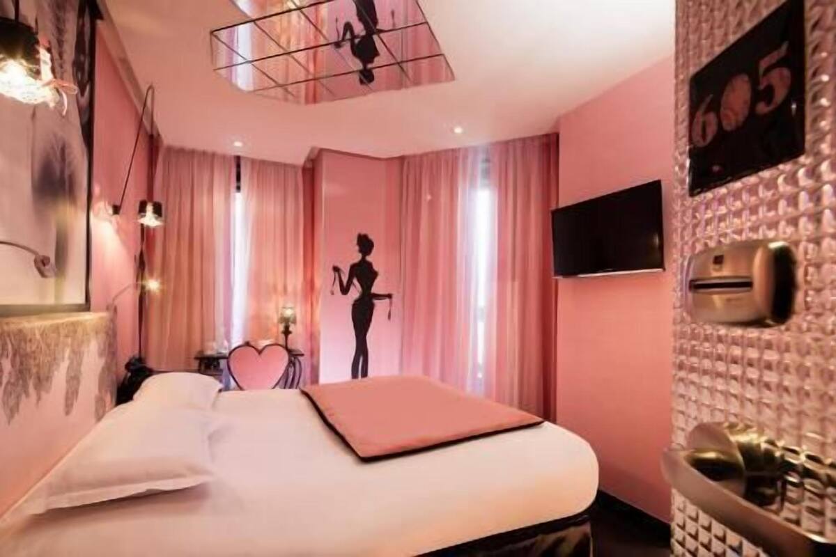Quarto de hotel com cama, televisão, minibar e uma grande janela com cortinas.