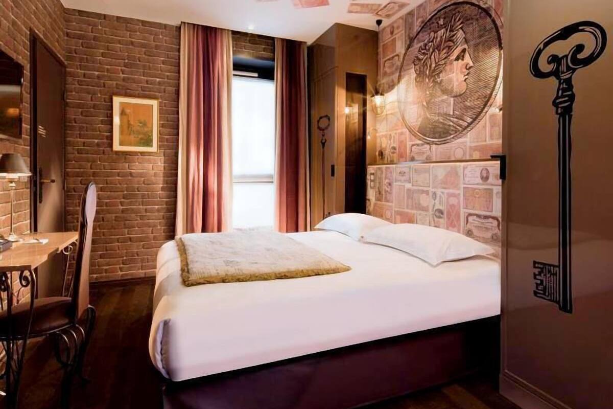 Quarto de hotel com parede de tijolos, cama com roupa branca, escrivaninha com cadeira, e parede com notas de dinheiro e um desenho de chave.