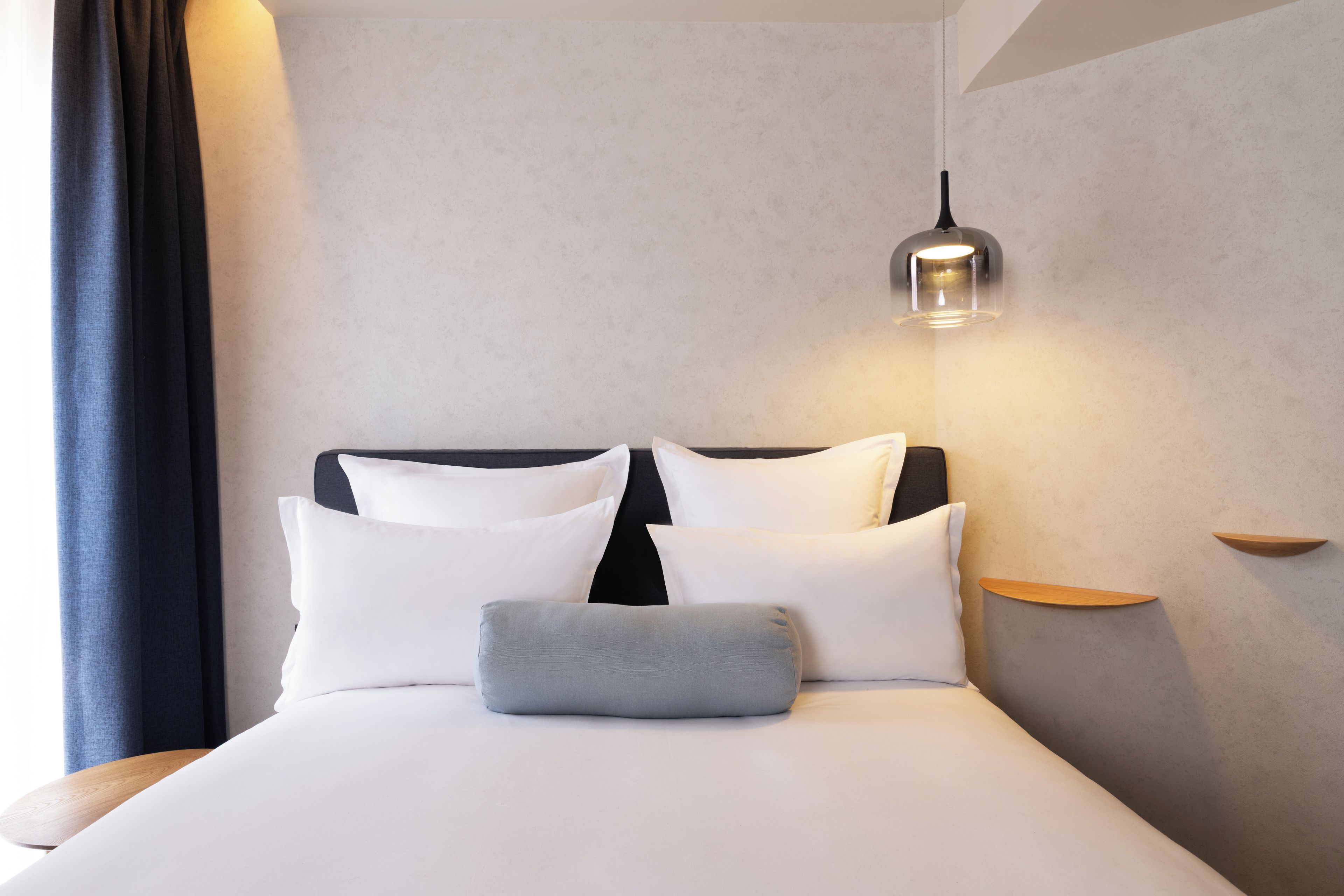 Een netjes opgemaakt bed met witte lakens en een grijs kussen, een zwart hoofdbord en een hanglamp erboven.