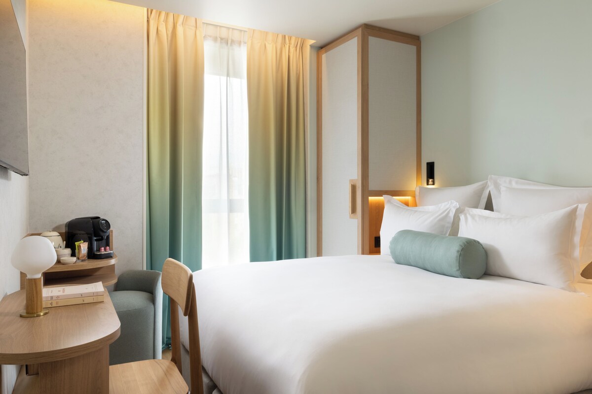 Habitación de hotel con cama, escritorio con cafetera, silla, armario y ventana con cortinas.