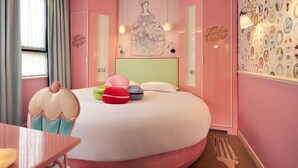 Ein zartrosa Schlafzimmer mit Bett, bunten Kissen, einem Nachttisch und einer Wand mit dekorativen Tellern.
