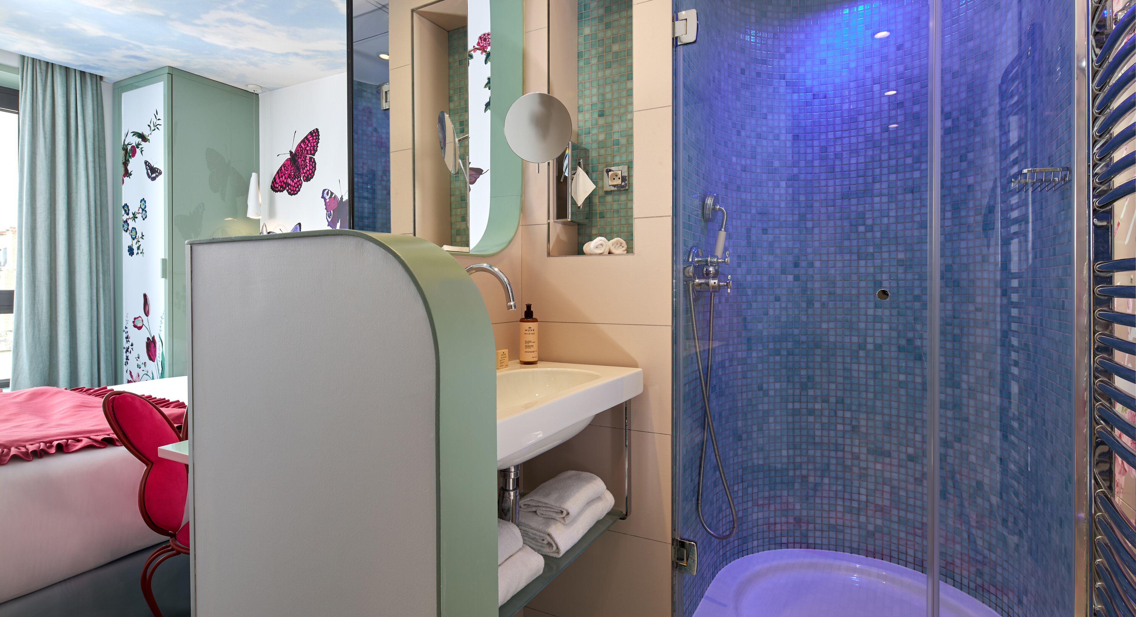 A bathroom with a shower, sink, and mirror. There is a bed with a red chair and a window with curtains.