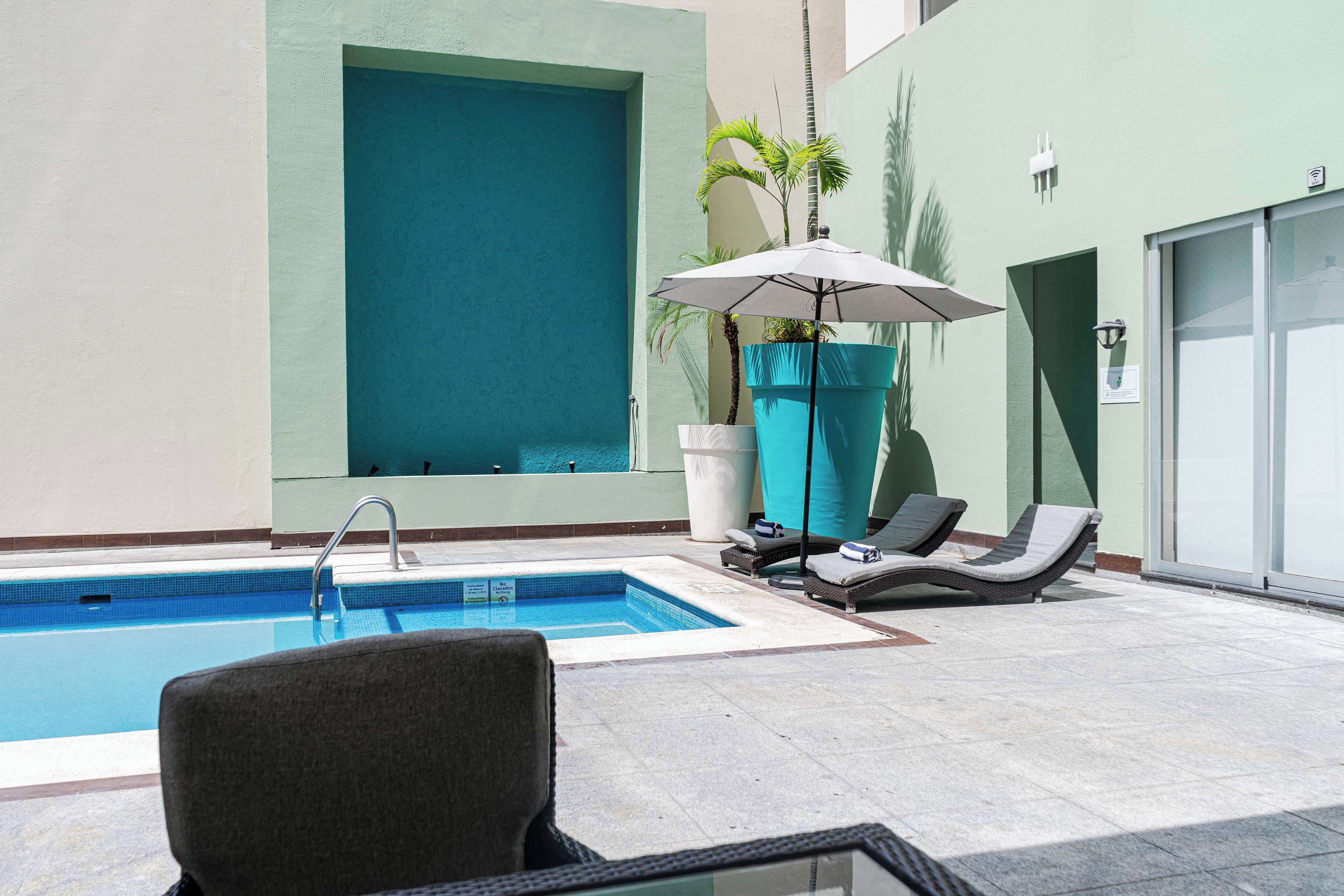 A pool area with lounge chairs and a large umbrella, a tiled pool, and a building with a green wall and a garage.