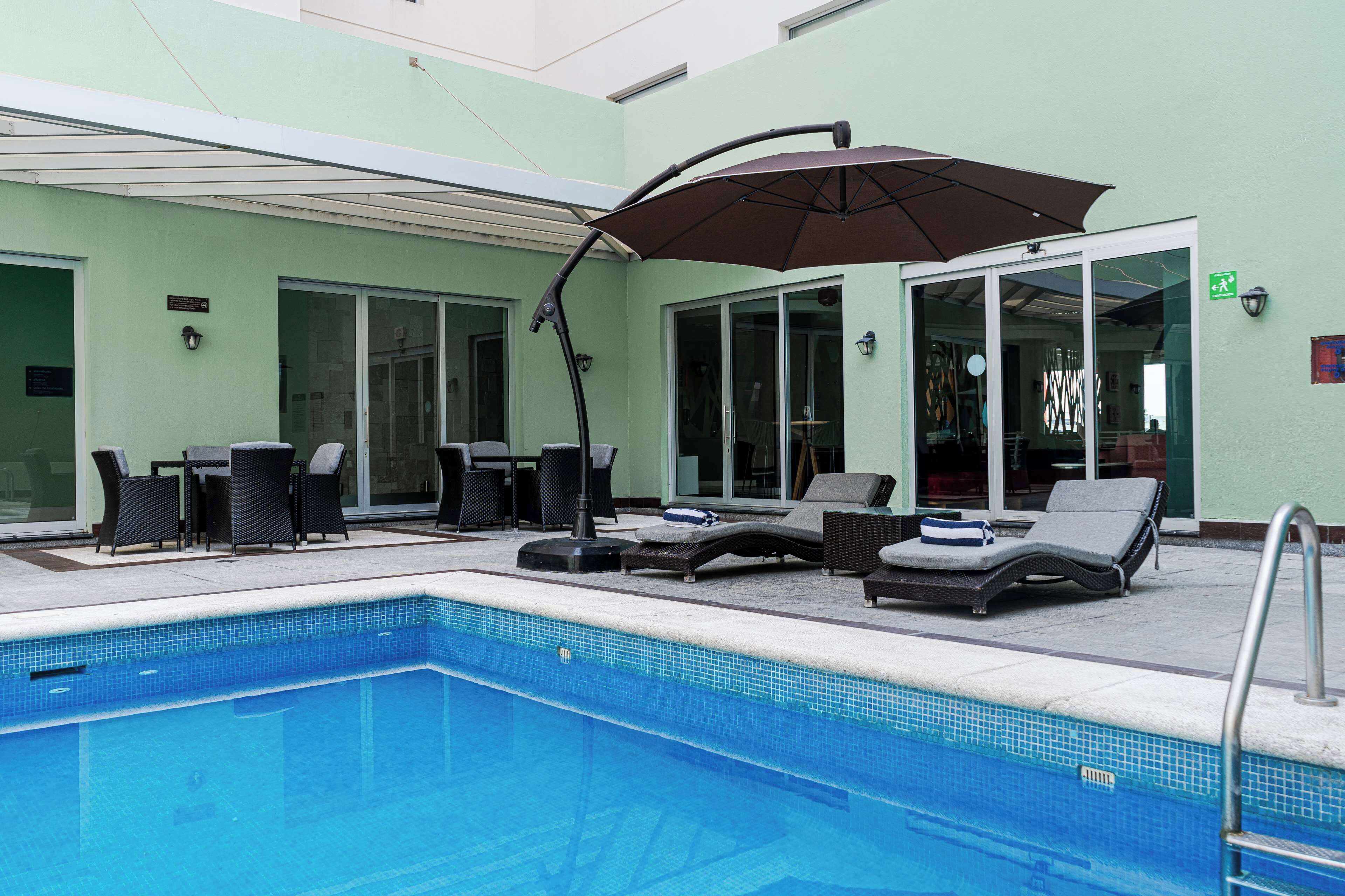 Una piscina con sillas de descanso y una mesa, rodeada por un edificio con puertas de vidrio y un muro verde.