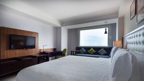 A modern hotel room with a large bed, a flat-screen TV mounted on the wall, a desk with a chair, and a window with a view of the sky.
