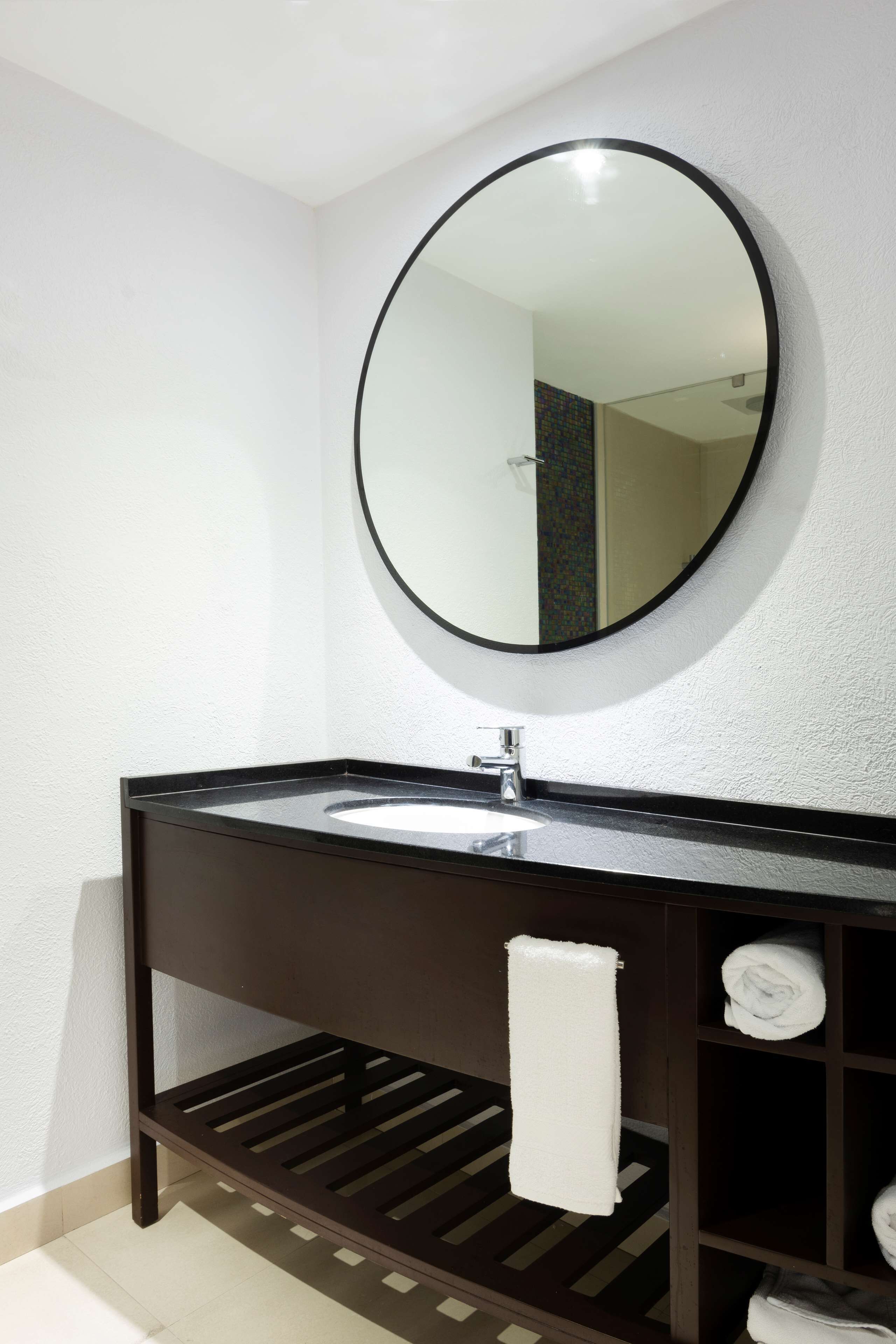 Un baño con un espejo redondo, un tocador de madera oscura, un lavamanos blanco y un perchero para toallas.