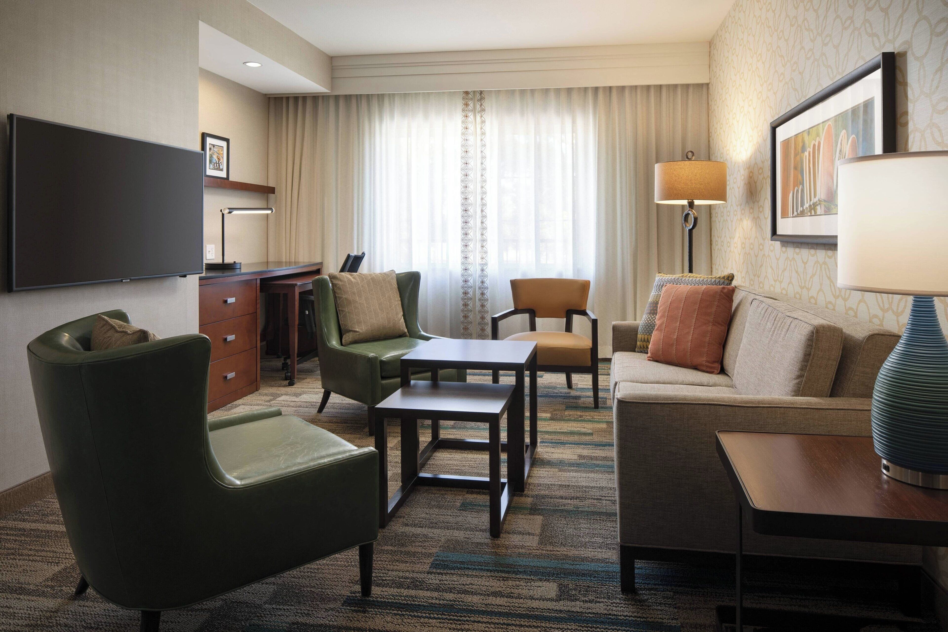 A hotel room with a sofa, armchairs, a small table, a TV, and a lamp.