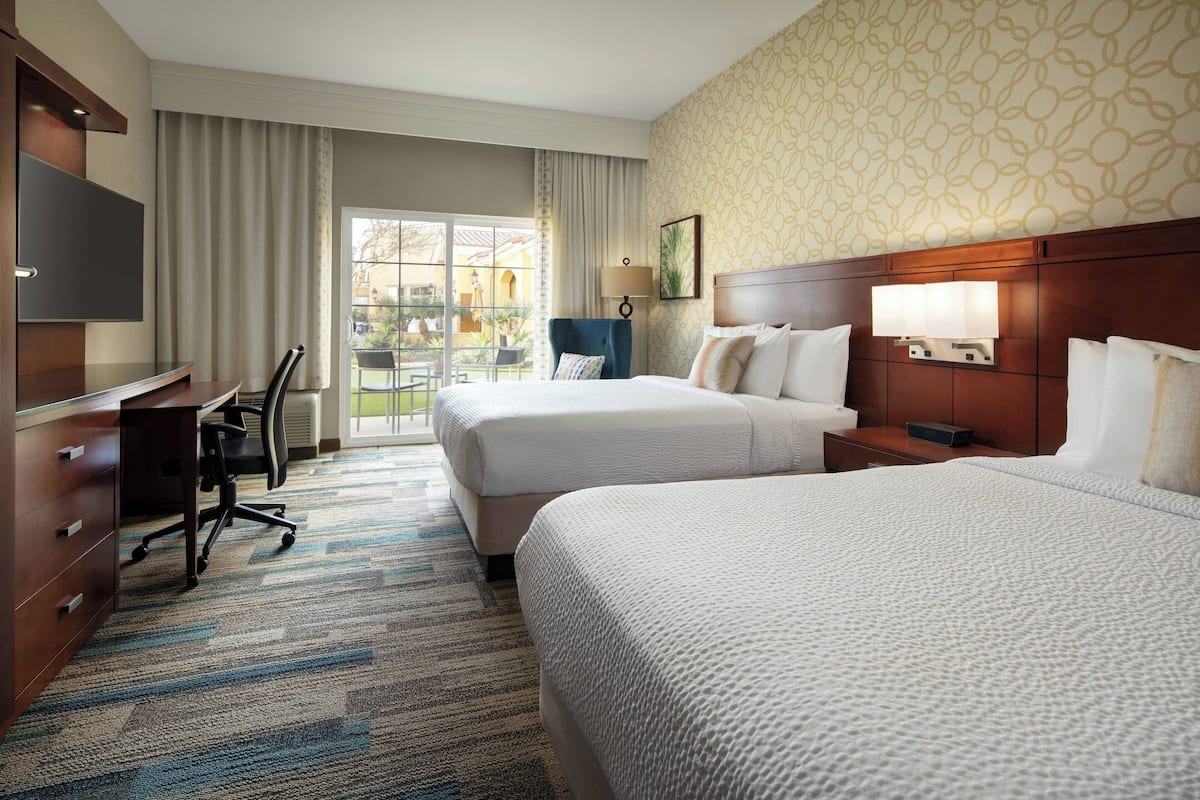 A hotel room with two beds, a desk, a chair, a TV, and a view of the outside.