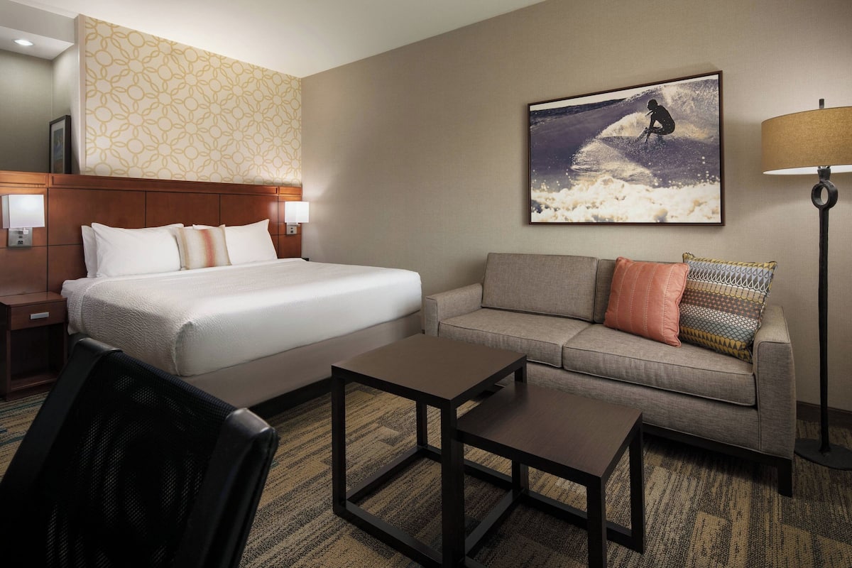 A hotel room with a bed, a sofa, a coffee table, a lamp, and a wall with a surfing-themed picture.
