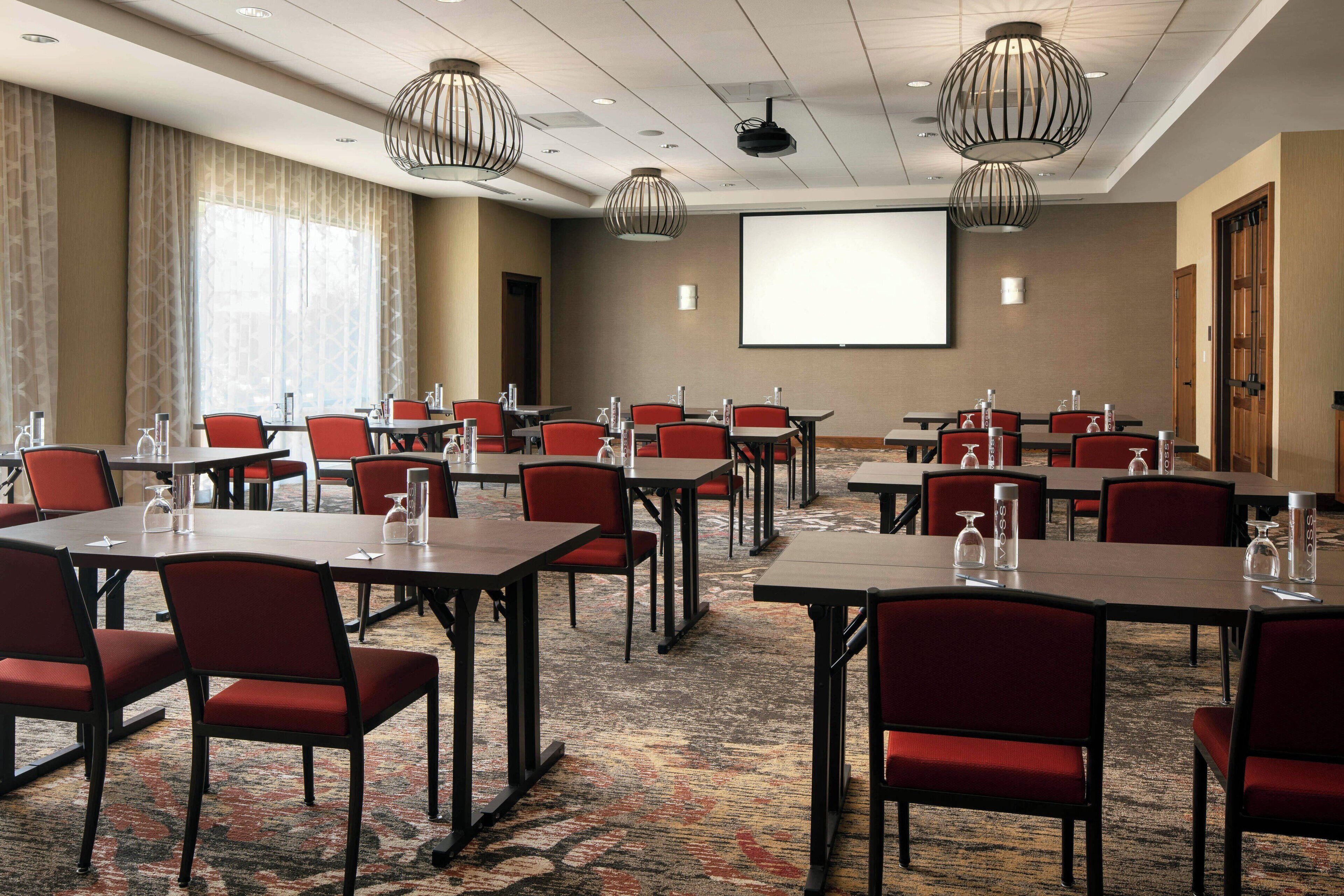 Une salle de conférence équipée de plusieurs tables et chaises, d’un écran de projection et d’une grande fenêtre avec des rideaux.