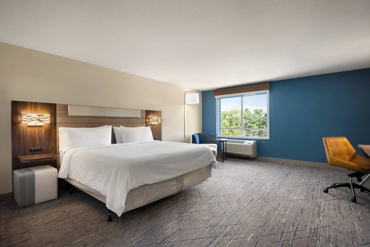 A hotel room with a large bed, a desk, a chair, a window with a view, and a wall-mounted air conditioner.