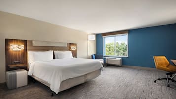 A hotel room with a large bed, a desk, a chair, a window with a view, and a wall-mounted air conditioner.