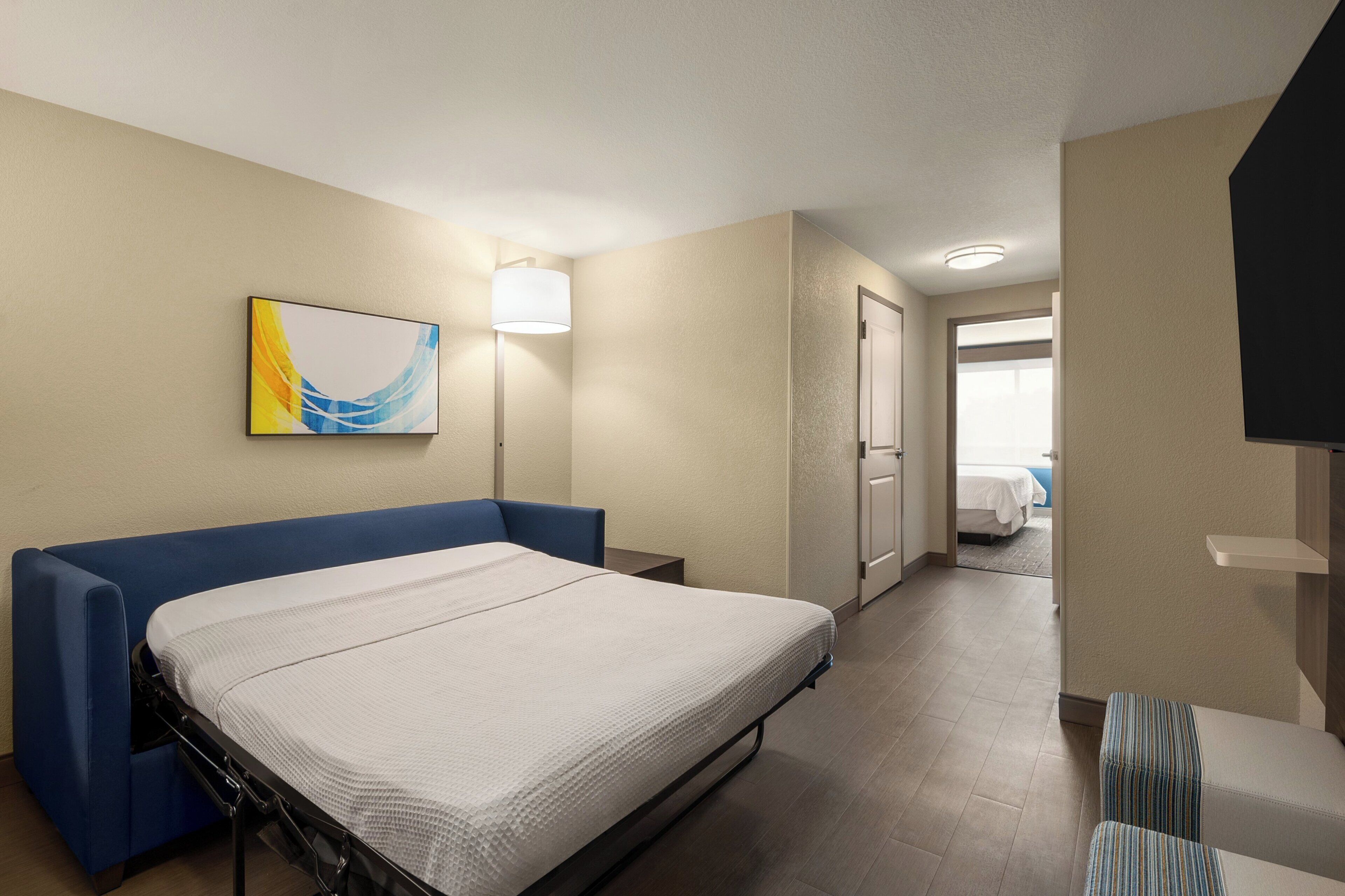 A hotel room with a bed, a desk, a chair, a TV, and a bathroom visible through an open door.