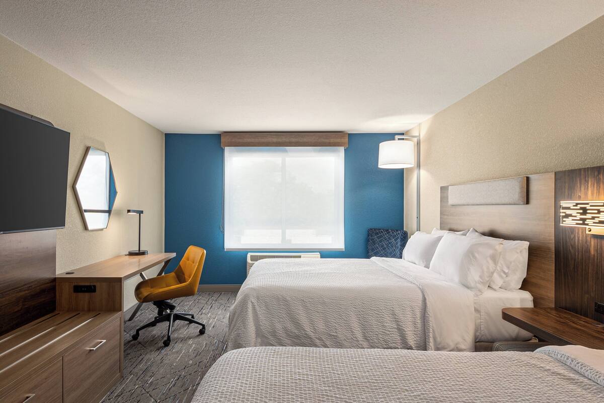 A hotel room with two beds, a desk, a chair, a TV, and a large window.