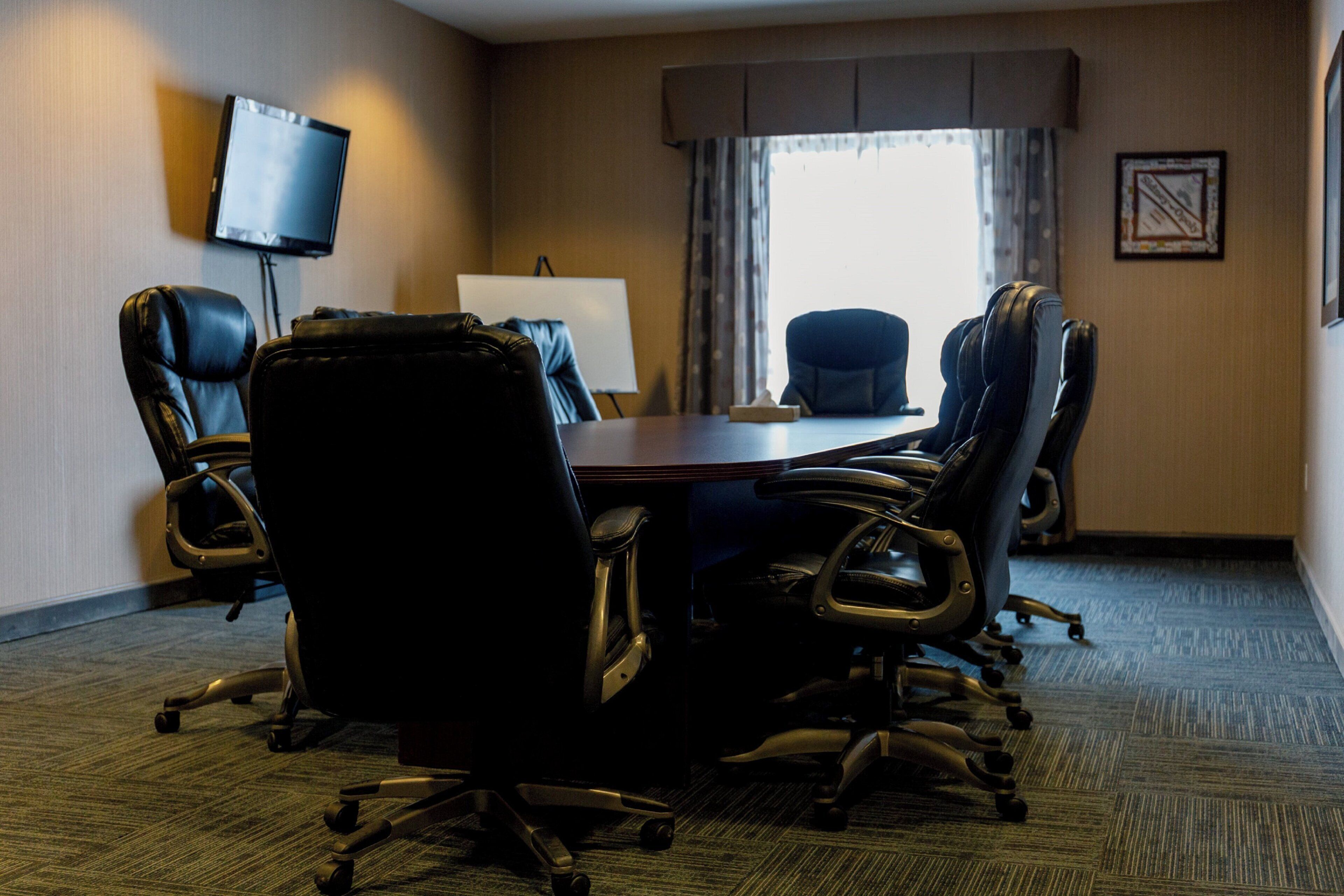 Une salle de conférence équipée d’une table ronde, de six chaises de bureau noires, d’un tableau blanc, d’un téléviseur à écran plat et d’une fenêtre avec des rideaux.