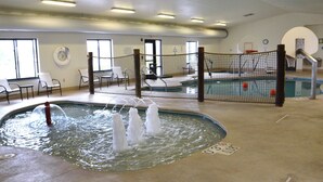 An indoor swimming pool with a water fountain feature, surrounded by a safety net, and a seating area with chairs.