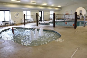 An indoor swimming pool with a water fountain feature, surrounded by a safety net, and a seating area with chairs.