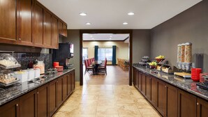 A modern kitchen with granite countertops, wooden cabinets, and a central island. There are various food items and utensils on the counters, and a dining area with chairs and a table in the background. - TownePlace Suites Midland (Midland)