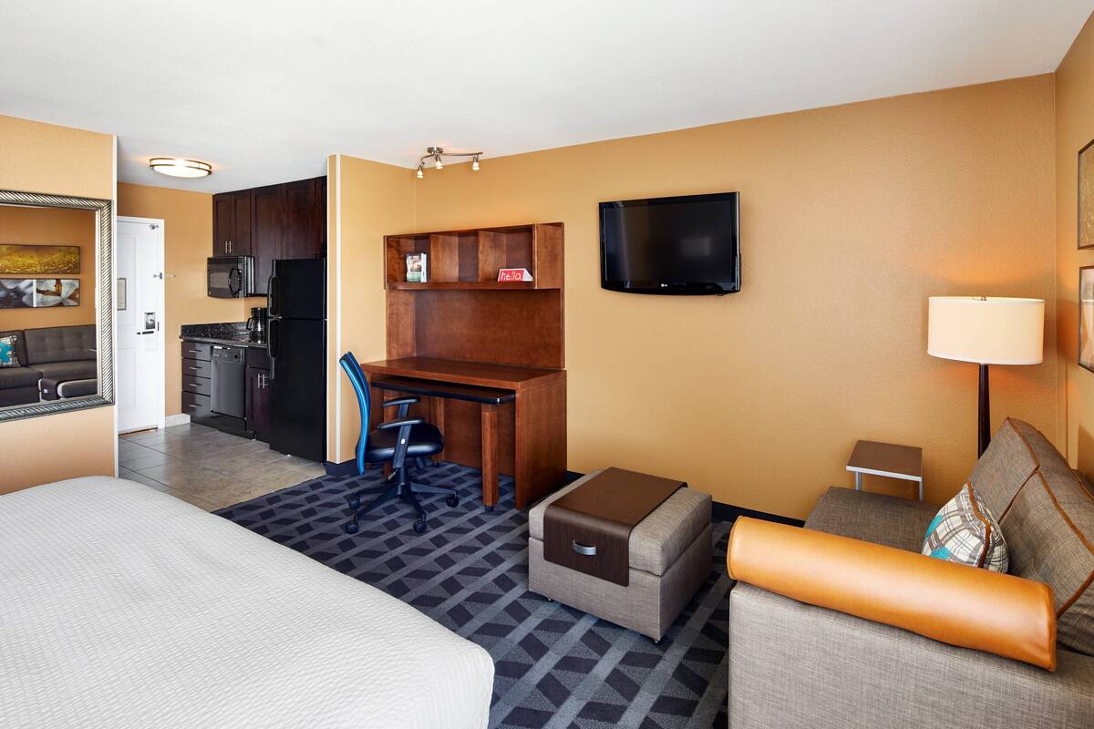A hotel room with a bed, a sofa, a desk with a chair, a kitchenette, a TV, and a lamp.