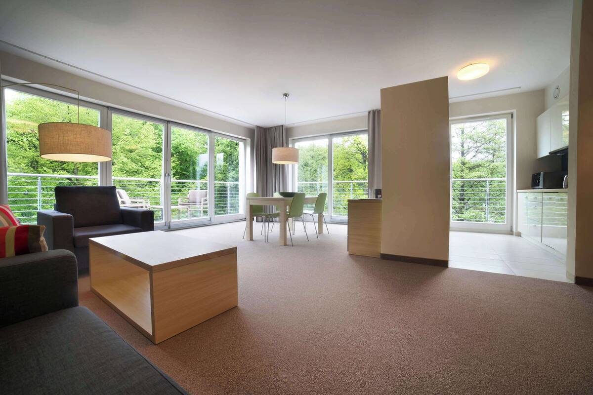 Un salon moderne avec un canapé, une table basse et un coin repas avec des chaises. De grandes fenêtres offrent une vue sur la verdure environnante.