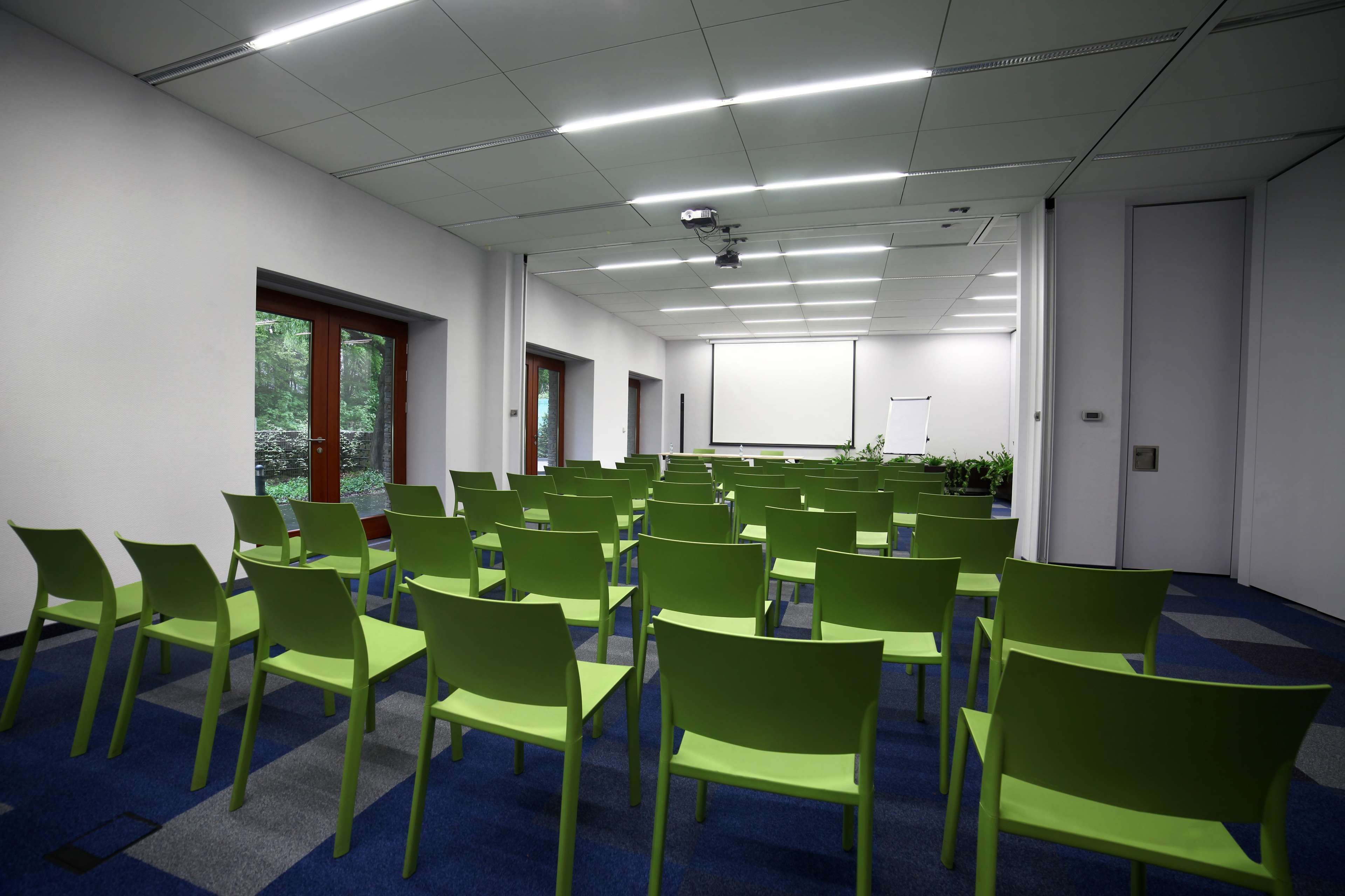 Un salón de conferencias con filas de sillas verdes, una pantalla de proyector y un amplio ventanal con vista a árboles.