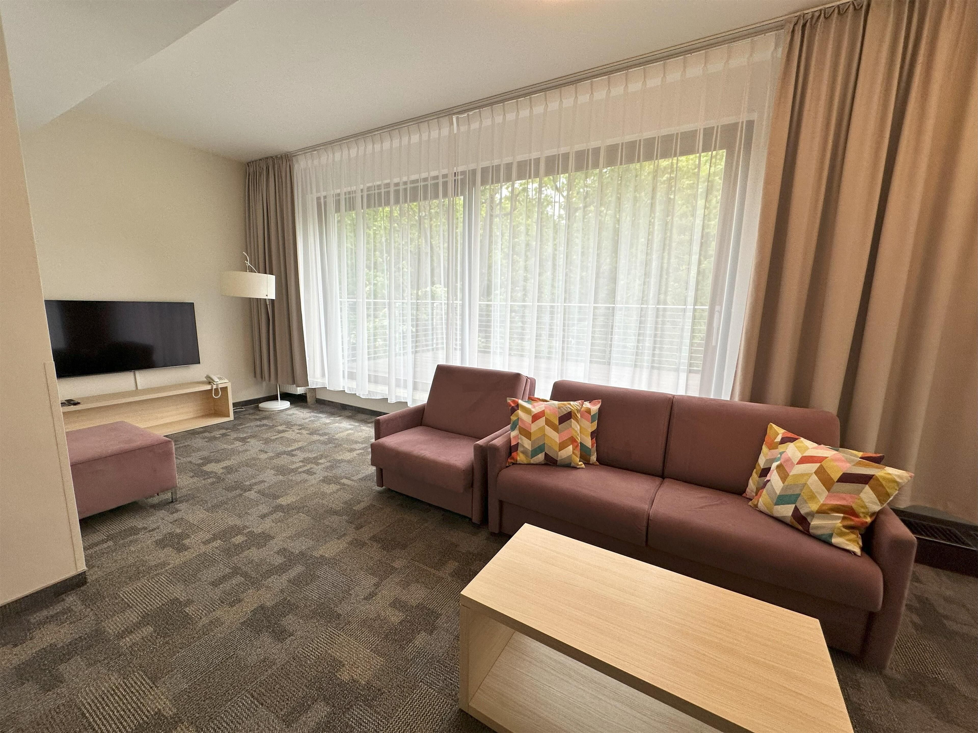 A modern living room with a flat-screen TV, a sofa, a coffee table, and a large window with sheer curtains.