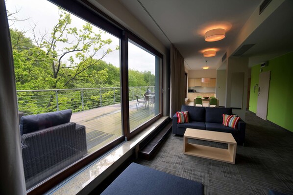 Una habitación de hotel moderna con ventanas amplias, un balcón, un sofá, una mesita de centro y una zona de cocina.