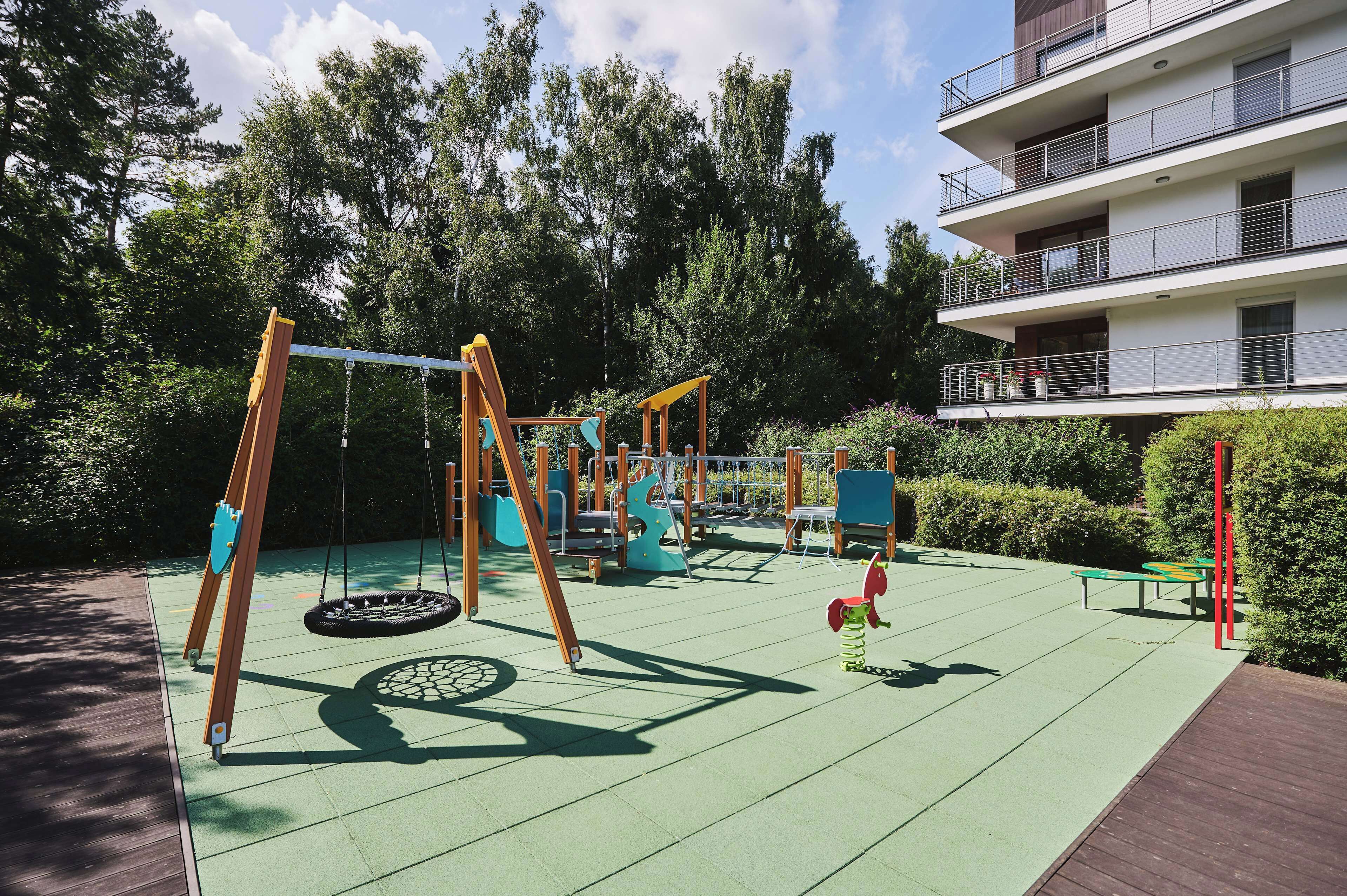 A playground with swings and a slide, surrounded by trees and a building.