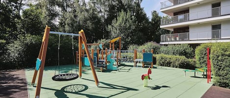 A playground with swings and a slide, surrounded by trees and a building.