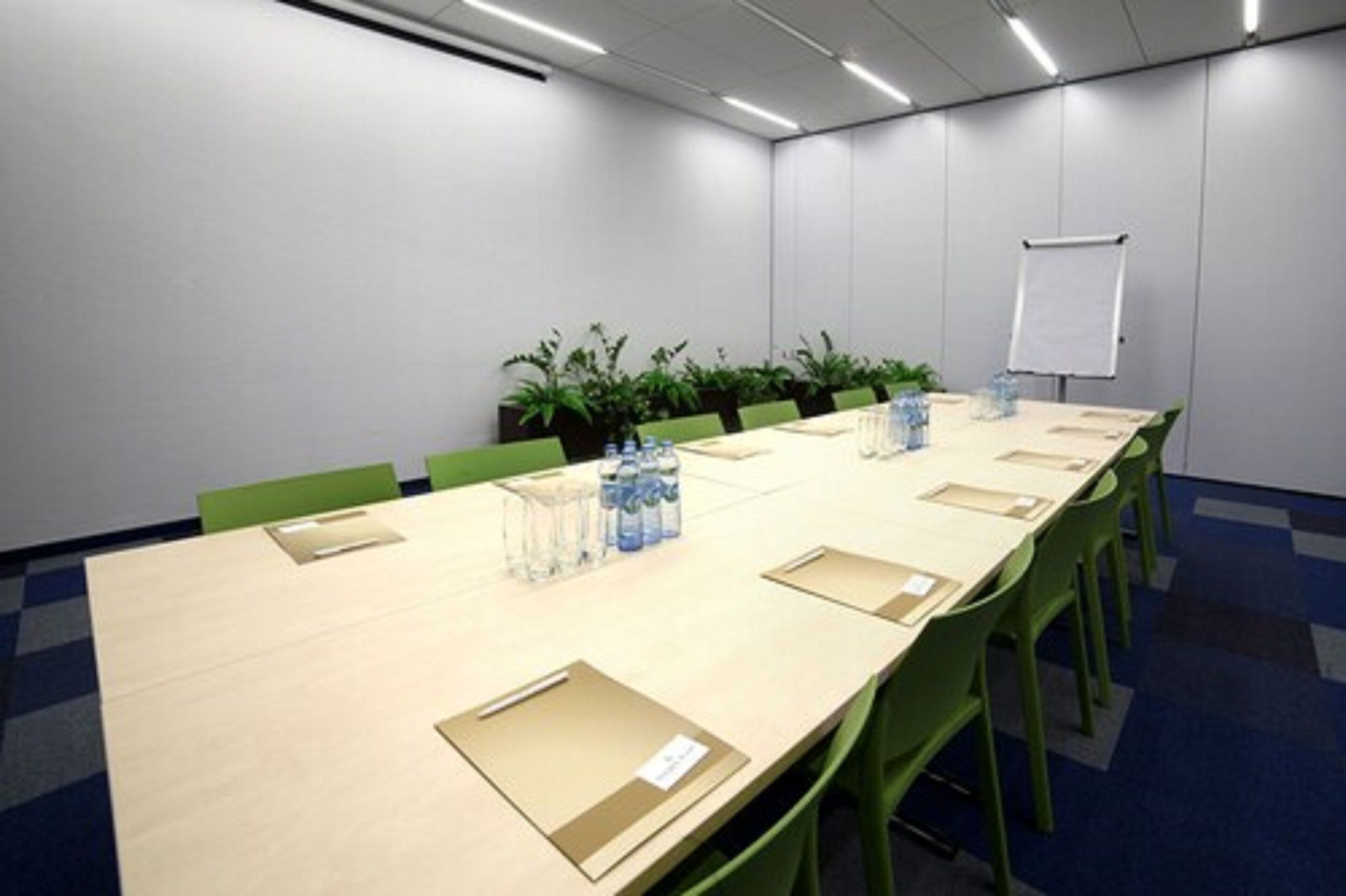 Sala de conferências com uma mesa comprida, cadeiras verdes, plantas em vasos, garrafas de água e um quadro branco.